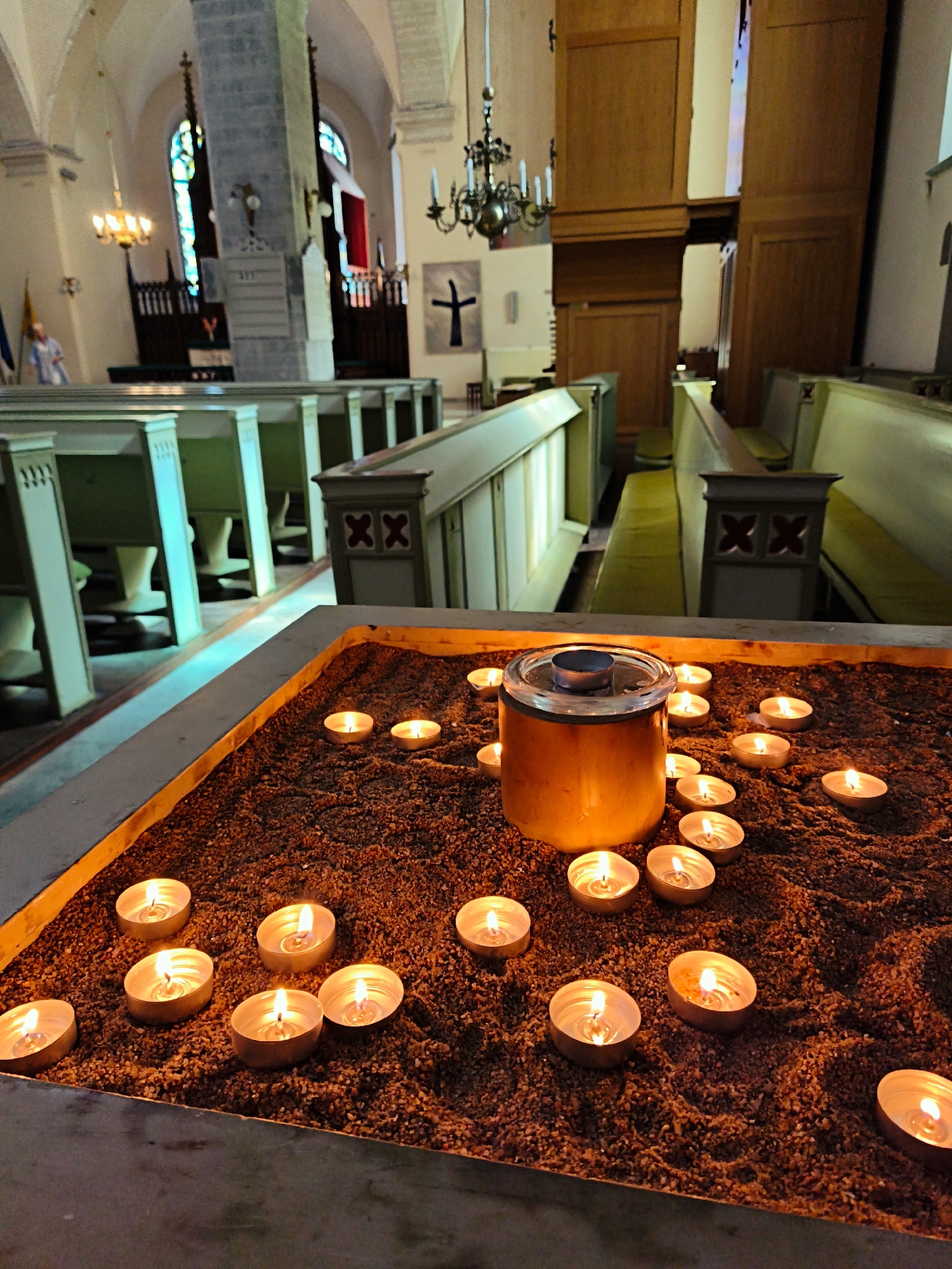 This is a close-up, low-angle interior shot of the interior of a church, likely located in Tallinn, Estonia, focusing on a sand-filled receptacle containing numerous lit tea light candles. The receptacle itself is a rectangular, shallow container with a light-colored rim. The sand appears to be a dark, granular material. In the center of the sand is a larger, cylindrical glass container with a metal lid, also lit from within.
Surrounding this central display are multiple small, lit tea light candles, each in a metallic holder, scattered across the sand. The flames of the candles create a warm, flickering glow, illuminating the sand and the immediate foreground.
In the background, the interior of a church is visible. Rows of dark green pews with lighter-colored accents are arranged in the nave. The architecture suggests a historic building, with arched windows and stone pillars. A chandelier hangs from the ceiling, and a large wooden structure, possibly an organ, is visible on the right. A simple black cross is mounted on the wall in the distance. The overall atmosphere is solemn and serene, typical of a place of worship.