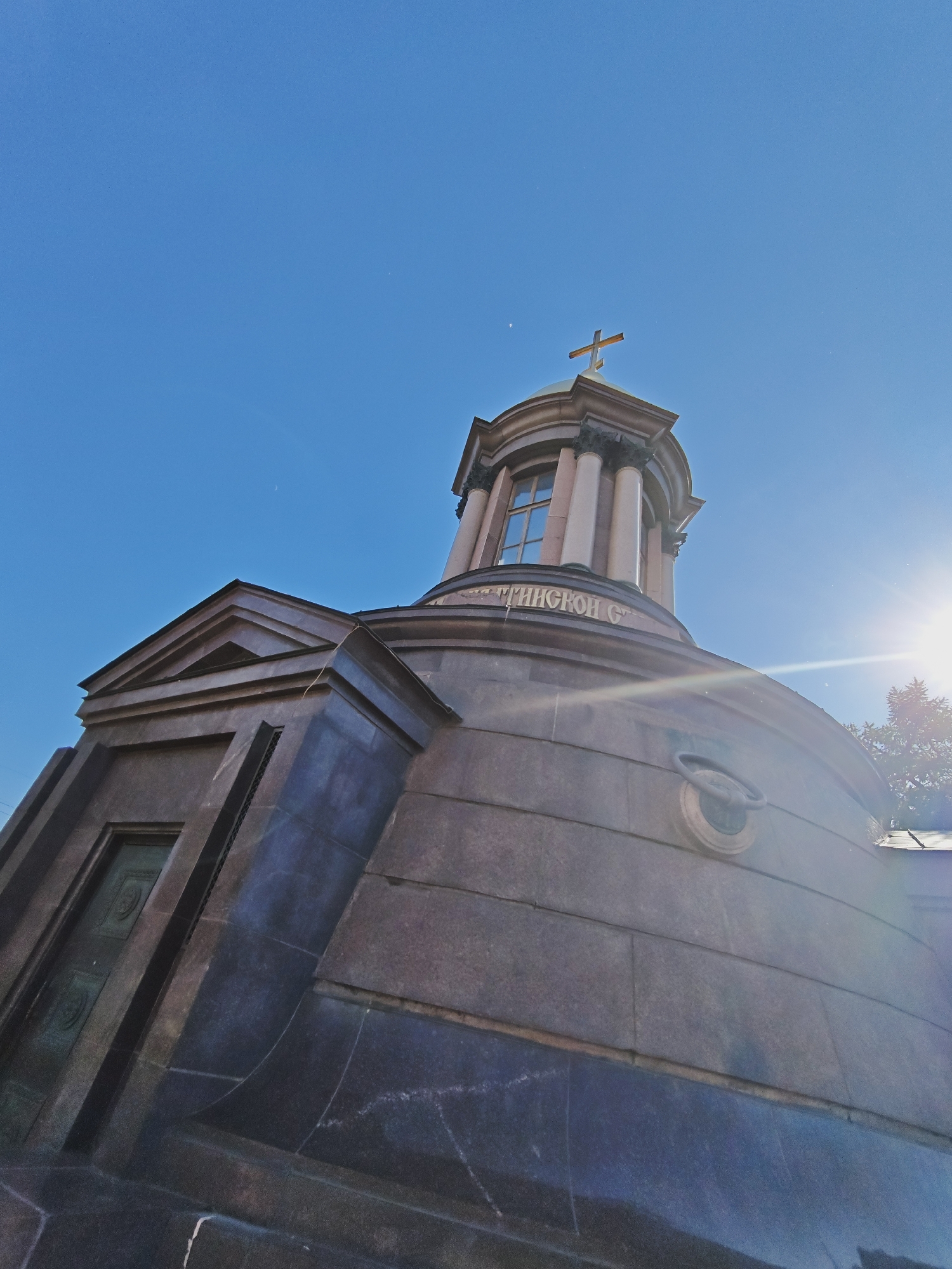 This image shows a low-angle shot of a large, ornate mausoleum or chapel in Saint Petersburg, Russia, set against a clear blue sky. The structure appears to be made of dark, polished granite, with classical architectural elements. A prominent feature is the domed rotunda at the top, supported by Corinthian columns and topped with a golden Orthodox cross. A circular window with multiple panes is visible on the rotunda.
To the left of the rotunda, a triangular pediment caps a section of the building with a recessed dark door that has ornate relief carvings. The building's curved facade is visible in the foreground, with a circular decorative element and a metal ring mounted on it. The sun is shining brightly, creating a lens flare and casting a warm glow on the scene, suggesting it is midday.
Visible text on the curved facade reads "КИНГСКОЙ С.", which appears to be part of a longer inscription. The overall impression is one of grandeur and historical significance, typical of the architecture found in Saint Petersburg. The clear sky and bright sun indicate fair weather. The image captures the imposing and majestic presence of the structure.