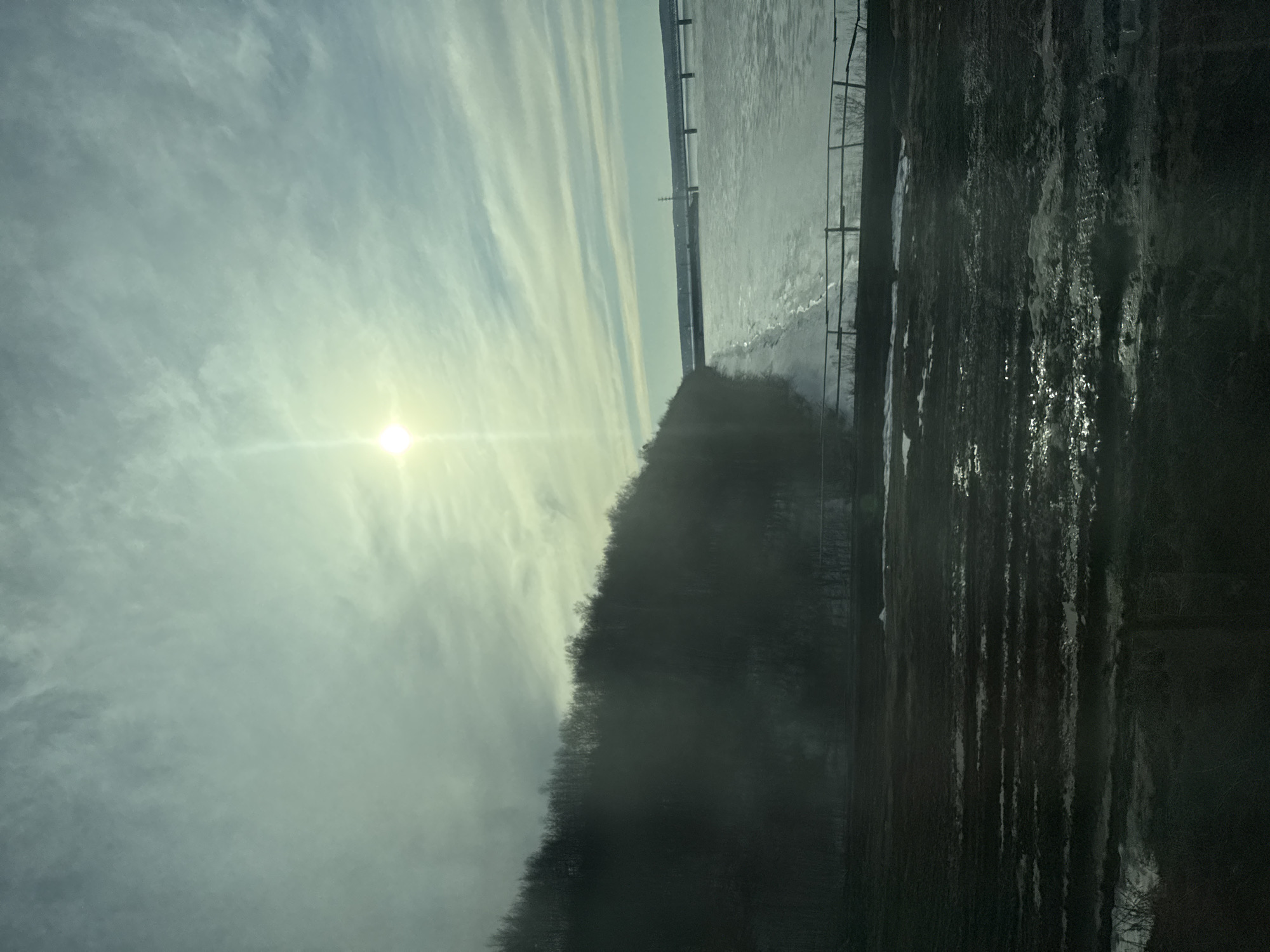 The image shows a landscape with a frozen river, a distant bridge structure, and a forested bank in the city of Perm, Russia. The sun is visible through a cloudy sky. A metal fence is also visible on the right side of the image. The foreground displays a dark, textured surface, possibly a wall or a muddy bank.