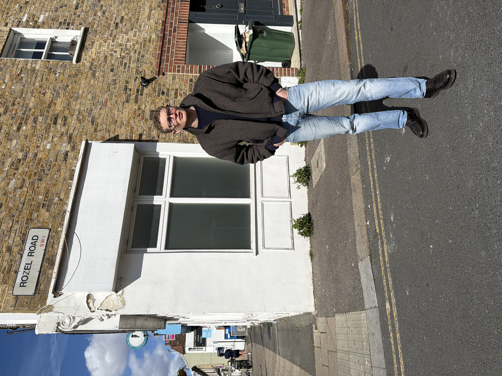 A person stands on a street, "Rozel Road SW4" in London, United Kingdom. The individual wears light-wash jeans, a brown jacket, and black boots. They are positioned on the asphalt, which features painted yellow double lines. Buildings line the street, including one with a white facade and a window. A street sign indicates "Rozel Road SW4". The weather appears to be sunny, and the sky is visible in the background.