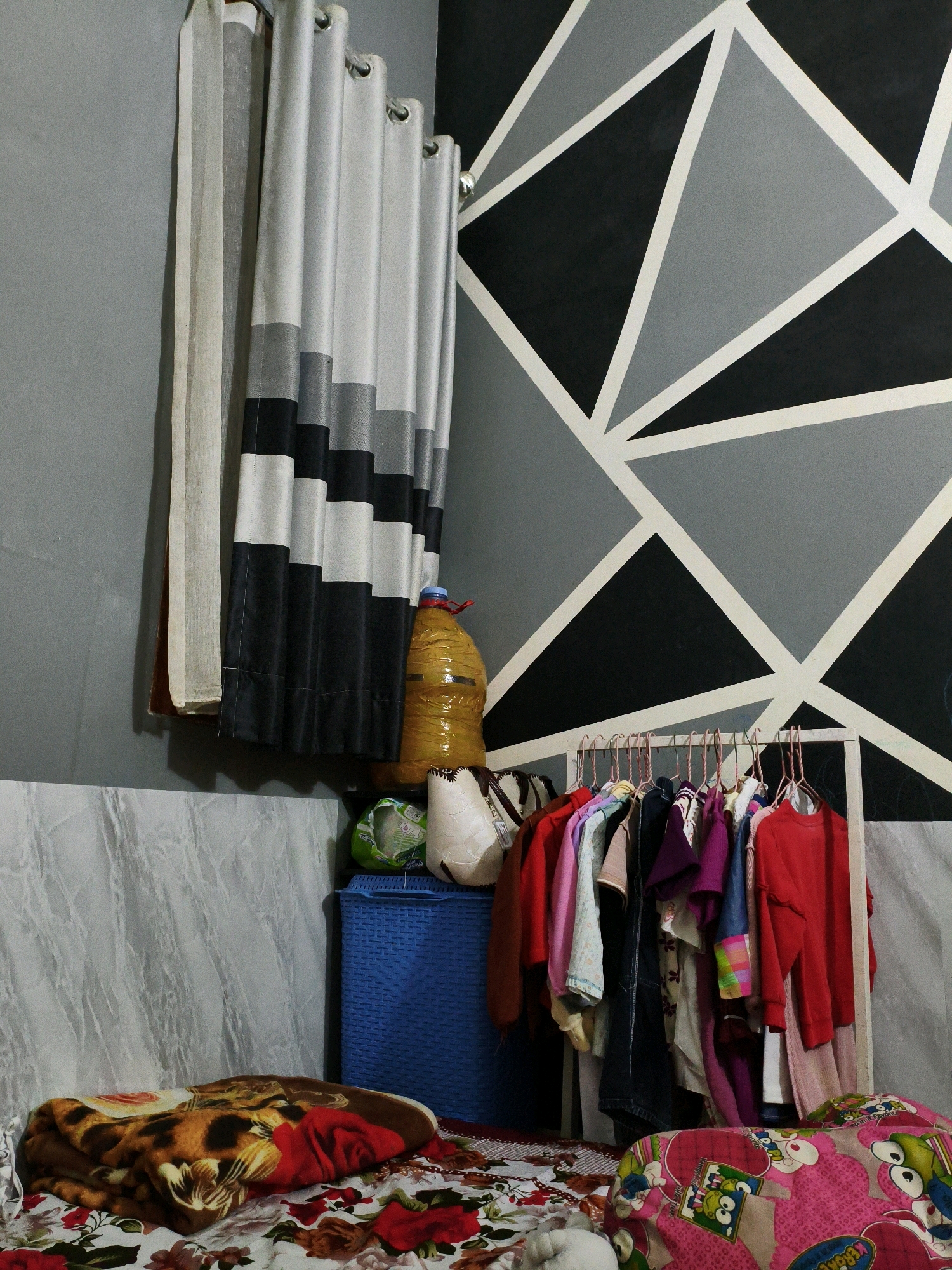 The image shows a cluttered bedroom scene in Karangtalun, Indonesia. A wall with a geometric pattern of black, gray, and white triangles forms the backdrop. To the left, a set of curtains hangs, one sheer and the others a striped pattern of black, gray, and white. In front of the wall, several objects are visible. A blue woven basket sits near the corner, topped with a yellow, bottle-shaped object wrapped in plastic and a green ball. To the right of the basket, a clothing rack is filled with a variety of colorful garments on hangers, including red shirts, pink shirts, and a denim jacket. A white frame surrounds the clothing rack. In the foreground, two beds are partially visible. One bed is covered with a blanket featuring a leopard print and red fabric. The other bed is covered with a pink blanket adorned with cartoon characters. The floor is covered with patterned fabric. No people are present in the scene.