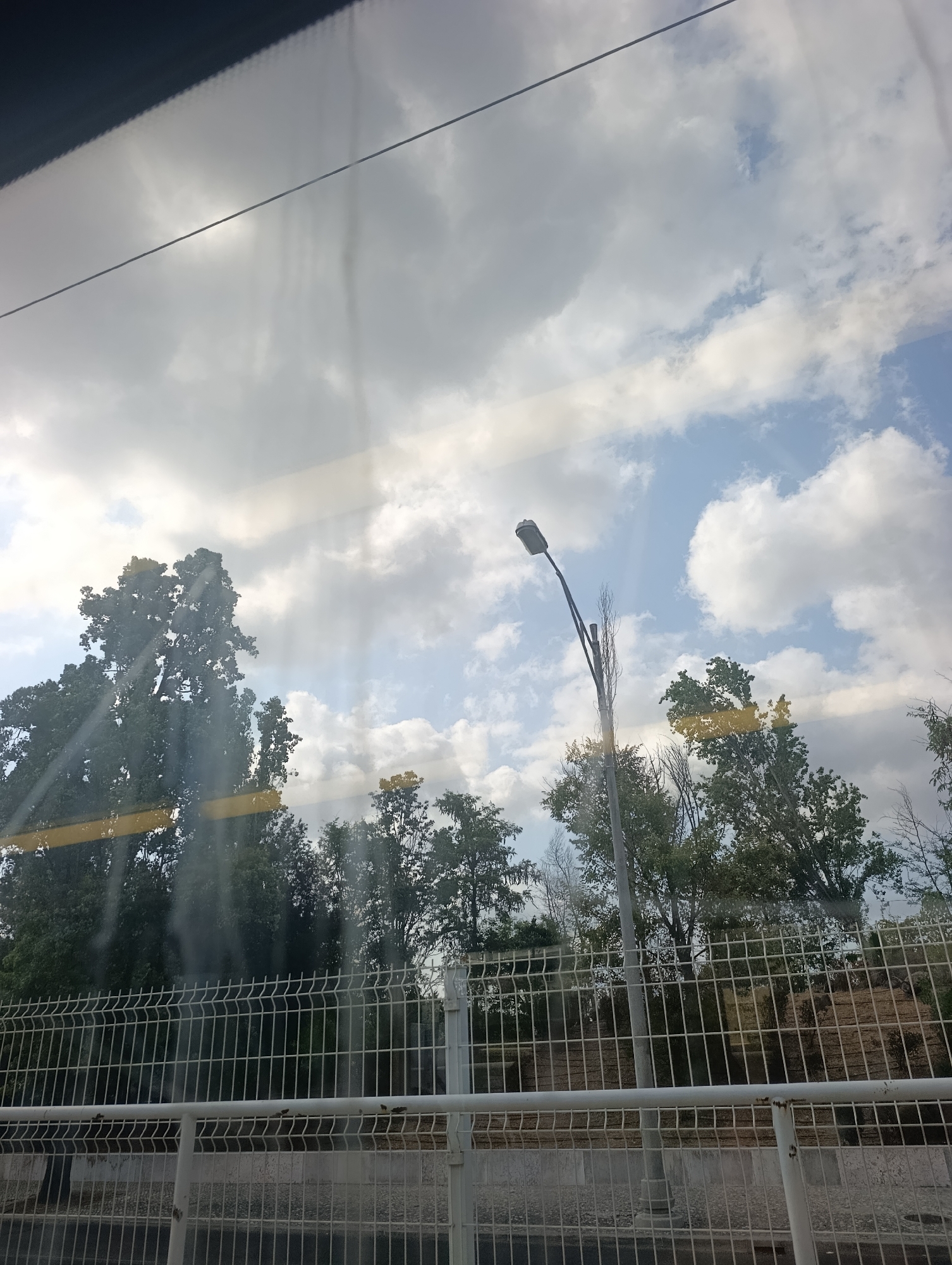 This is a photograph taken from inside a vehicle, likely a bus or train, looking out through a window. The window has visible reflections and streaks, particularly a horizontal yellow streak across the middle, and a dark, jagged line at the top.
The scene depicted is an outdoor environment. In the foreground, a white metal fence with diamond-shaped openings runs horizontally across the lower portion of the image. Behind the fence, there is a raised embankment with sparse vegetation and some scattered rocks. Further back, a line of trees with dense green foliage is visible.
Dominating the mid-ground and extending into the upper half of the image are several tall trees. The tree on the far left is particularly large and has a full canopy of dark green leaves. To its right, a tall, slender lamppost with a single light fixture at the top is positioned. Beyond the trees, the sky is visible, filled with a mix of white and gray clouds against patches of blue sky. The lighting suggests it is daytime, possibly late afternoon or early evening, with the sun creating some glare on the window.
There are no people visible in the image. The context of the photograph, a view through a vehicle window with reflections, suggests a journey. The specific location of Lisbon, Portugal, is not discernible from the visual elements present in the image itself; this information would need to be provided externally.