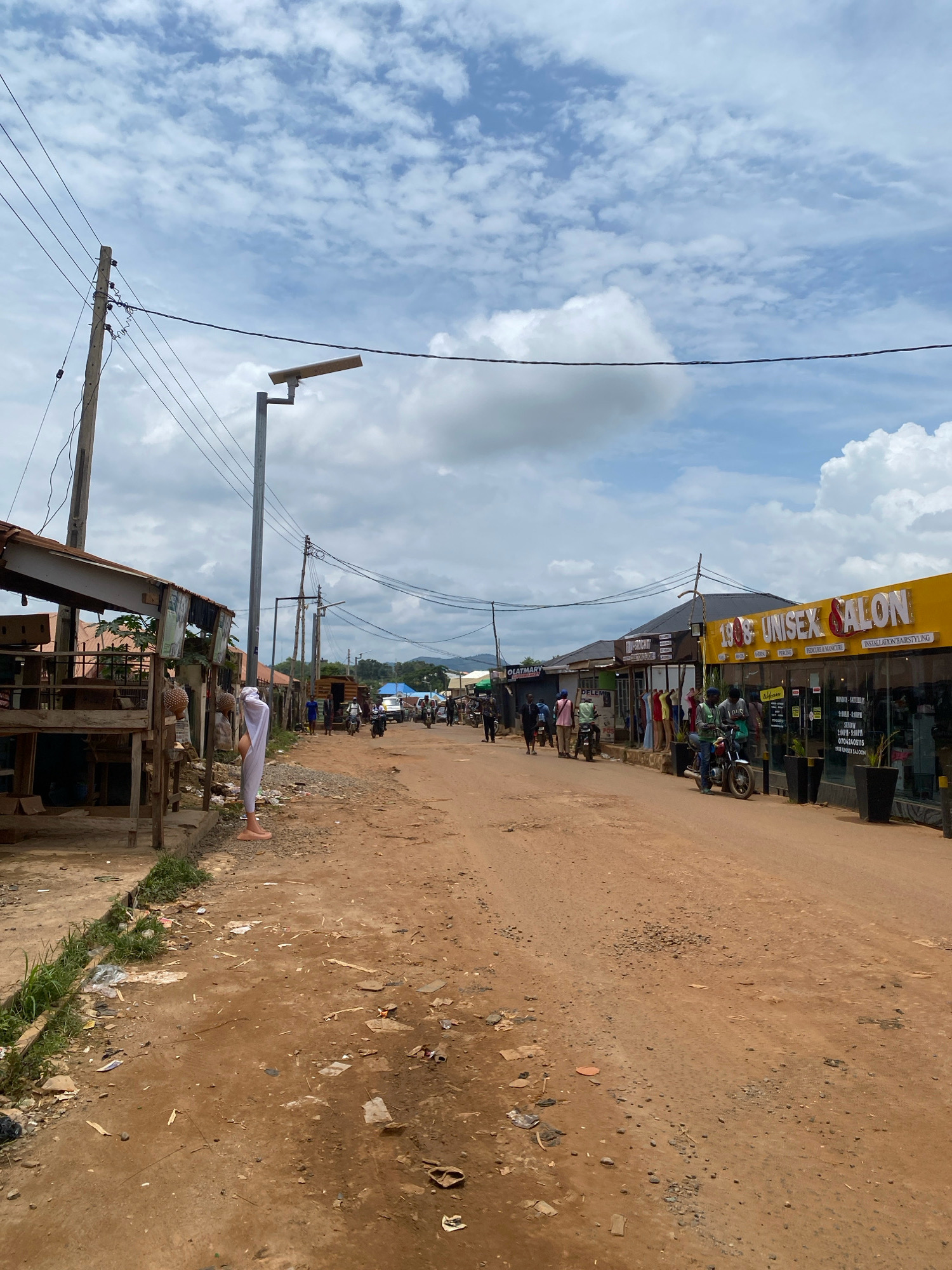 The image depicts a street scene in Iworoko, Nigeria. The unpaved road is reddish-brown, with scattered debris and some small stones. On the left side of the road, there are open-air stalls with wooden structures and corrugated metal roofs. One stall features a mannequin or statue covered in white fabric, with bare feet visible. Further down the street, on the right, is a building with a bright yellow facade, identified by signage as "1908 UNISEX SALON". This establishment appears to be a commercial storefront with large glass windows. Several people are walking on the street and sidewalk, and some are on motorcycles or scooters. Power lines and utility poles are visible overhead, stretching across the sky, which is partly cloudy with patches of blue. The background shows more buildings and a distant landscape with hills under the cloudy sky. The overall impression is of a busy, working-class urban environment.