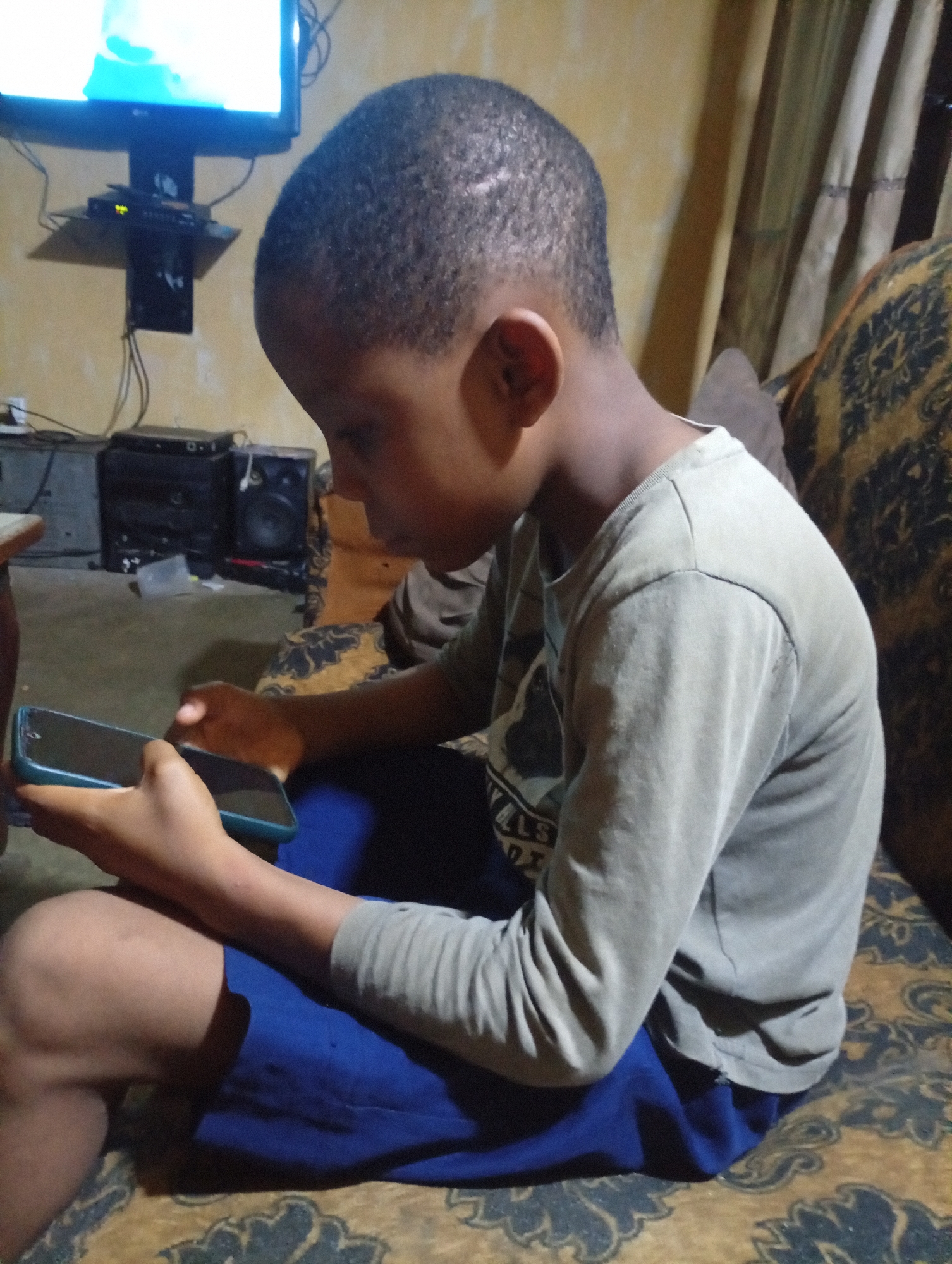 A young Black boy is seated on a patterned sofa, looking down at a smartphone he is holding. He is wearing a long-sleeved grey shirt and blue shorts. In the background, a television is mounted on a wall above a stereo system. The location appears to be inside a home in an unknown city.