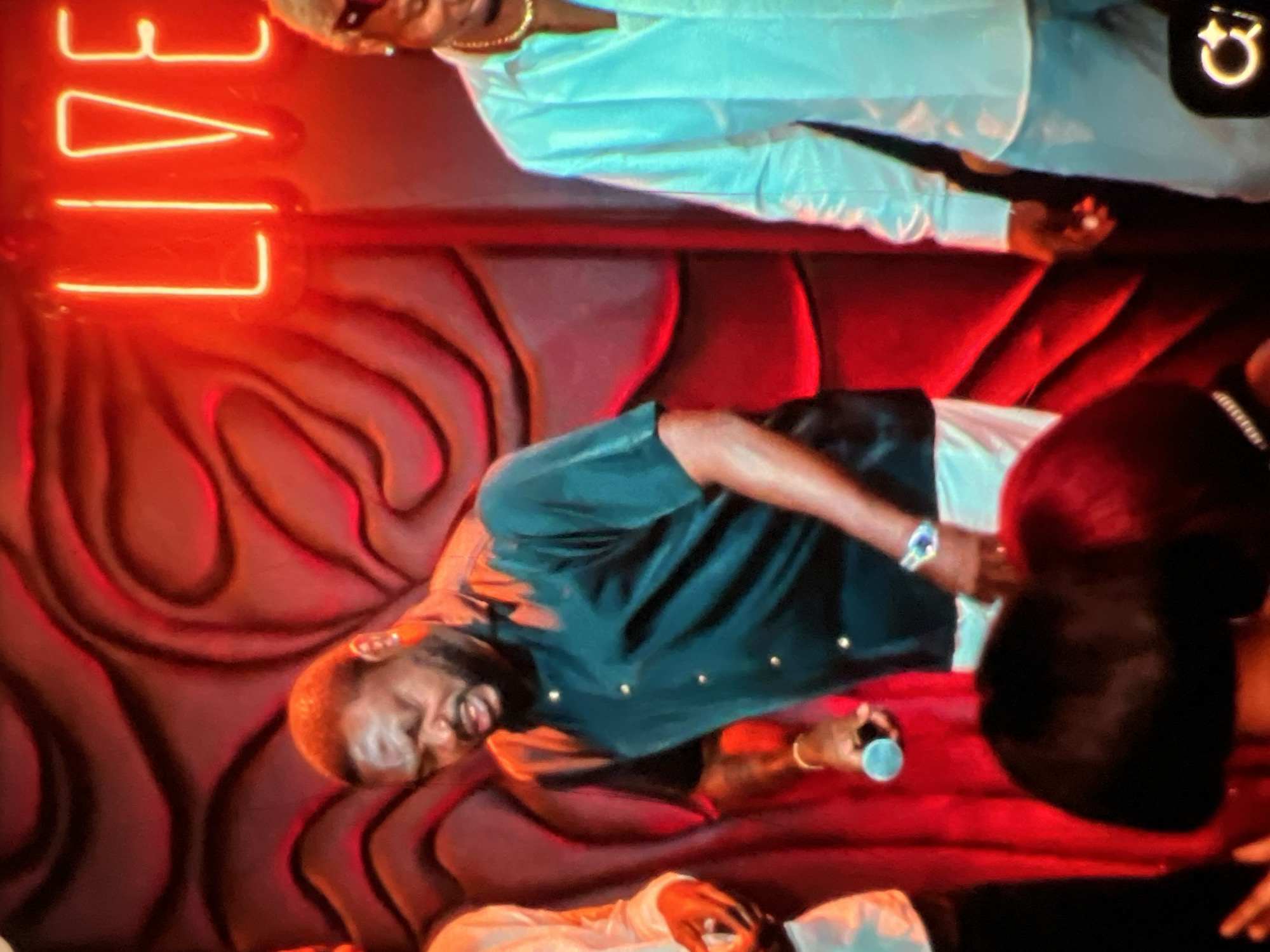The image depicts a live performance setting in Lagos, Nigeria, illuminated by red and neon lights. In the foreground, a man in a dark green short-sleeved shirt and white pants is holding a microphone, appearing to be singing or speaking. He has a beard and is wearing a wristwatch. To his right, another man in a light blue shirt and white pants is visible, wearing sunglasses and a gold chain. A neon "LIVE" sign is prominently displayed on the wall behind them, bathed in red light. The background features a textured red wall with an organic, wave-like pattern. In the bottom portion of the image, the heads of audience members are visible, with dark hair illuminated by the stage lights. The overall atmosphere suggests a vibrant nightlife or entertainment venue.
