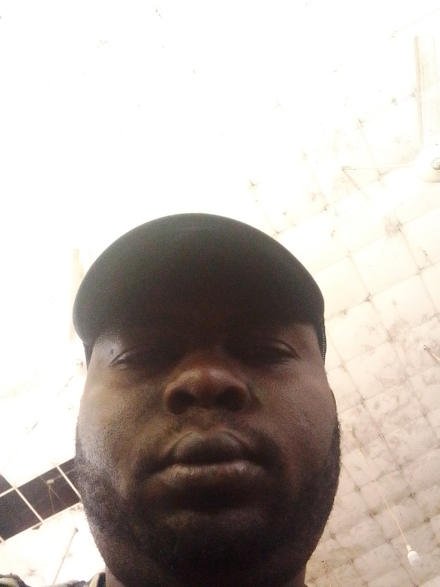 A close-up, low-angle selfie shows the face of a Black man wearing a dark baseball cap. The man's head is angled upwards, filling most of the frame. His eyes are visible, and he has stubble on his chin and cheeks. The background appears to be a ceiling with a tiled or patterned surface, and a light fixture is visible in the upper right corner. The overall lighting is bright, suggesting an indoor environment. The image does not provide information about a specific city or location.