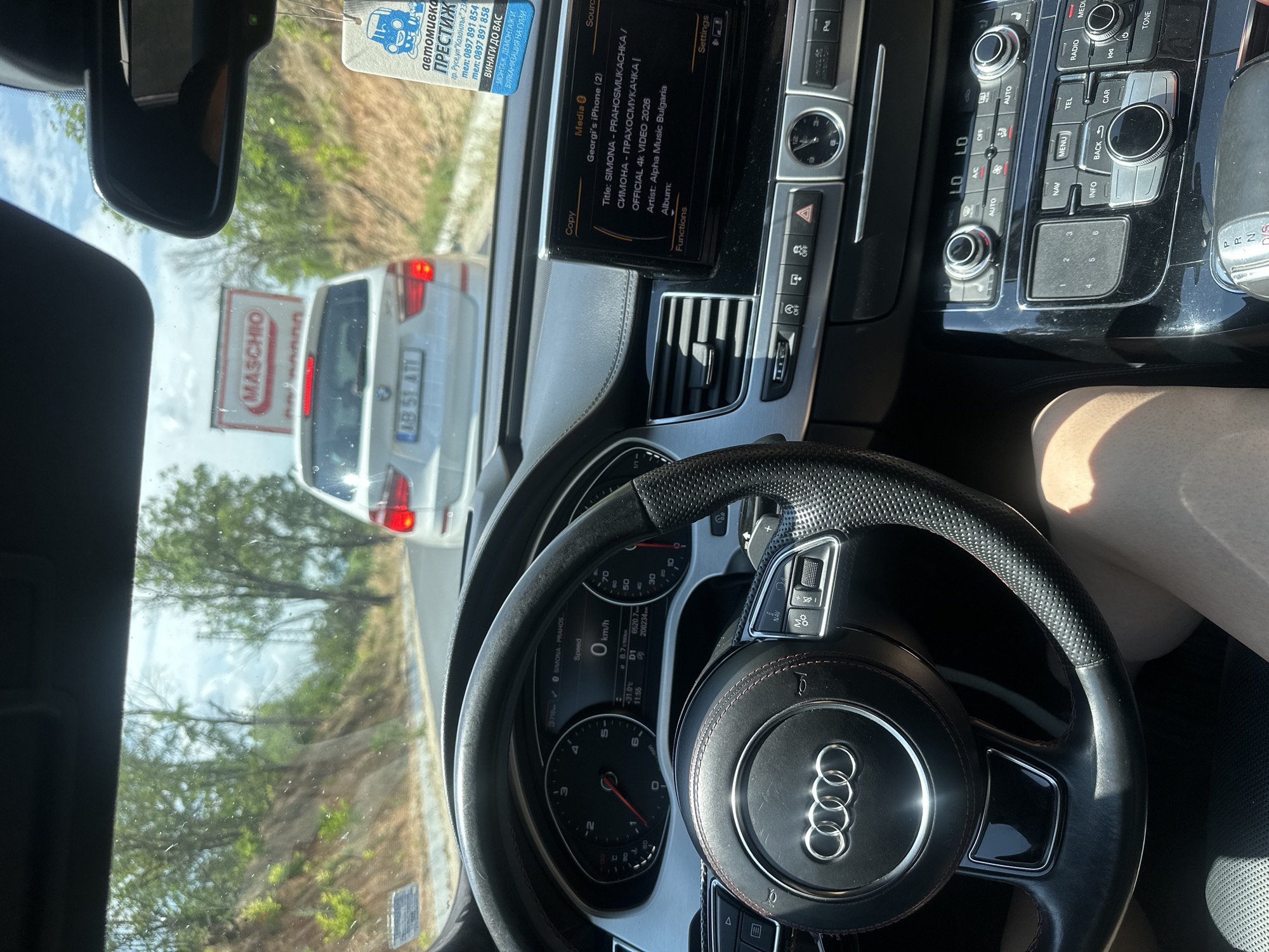 The image shows the interior of a car, likely an Audi, as indicated by the steering wheel emblem and dashboard controls. The driver's perspective is evident, with the steering wheel, dashboard, and infotainment screen prominently featured. Outside the car, through the windshield, a white van with license plate "AB 51 ATI" is visible, suggesting the car is in traffic. The setting appears to be a sunny day, with trees and a roadside embankment visible through the windows.
On the infotainment screen, song information is displayed, identifying the media as "SIMONA - PRAHOSMUKACHKA / СИМОНА - ПРАХОСМУКАЧКА | OFFICIAL 4K VIDEO 2026" by "Alpha Music Bulgaria," streamed from "Georgi's iPhone (2)."
A tag hanging from the rearview mirror displays text in Bulgarian, including the name "ПРЕСТИЖ" (Prestige), a car wash service located in Ruse, Bulgaria, with contact phone numbers. Another tag has the word "MASCHIO" on it.
The dashboard indicates the car is currently stationary, with the speed reading "0 km/h." The time shown is "11:55." The overall mood is relaxed, possibly indicating a stop or slow traffic during a journey. The presence of the car wash advertisement and the Bulgarian text suggests the location is in Bulgaria.