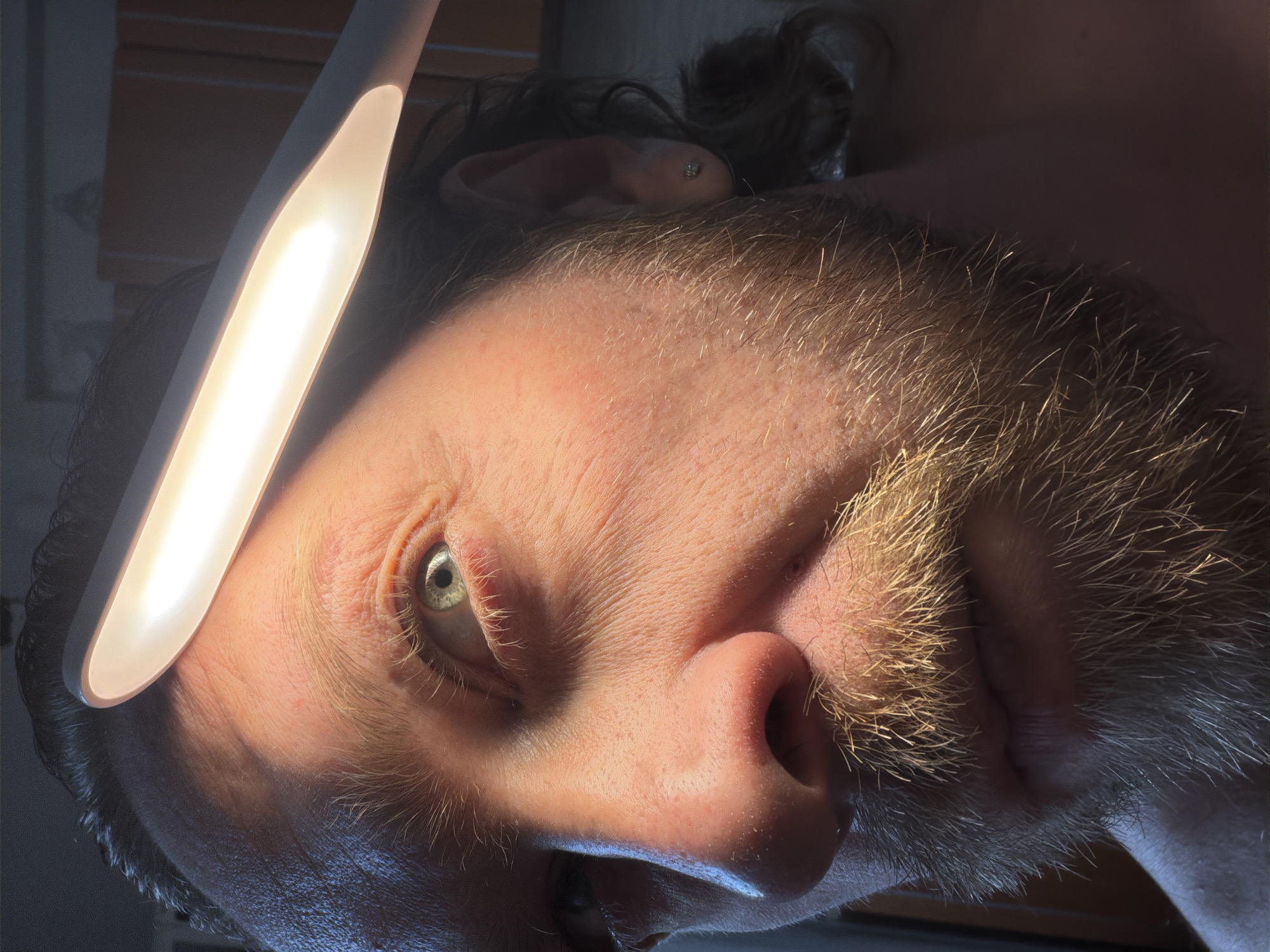 A close-up, high-contrast image captures the right side of an adult male's face, illuminated by a linear, warm white LED light source positioned diagonally above his forehead. The individual has light-colored skin, a prominent light brown/blonde beard and mustache, and dark hair. His right eye, light blue or green, is open and appears to look slightly upwards and to the right. Fine wrinkles are visible around the eye and on the forehead. A small, silver-colored stud earring is present in the right earlobe. The strong overhead light casts a distinct shadow across the left side of the face, partially obscuring the nose and mouth area. The background is dark and out of focus, revealing faint outlines of what appear to be shelves or cabinets, indicating an indoor environment. No visual information within the image allows for corroboration of a specific location in Saint Petersburg, Russia.