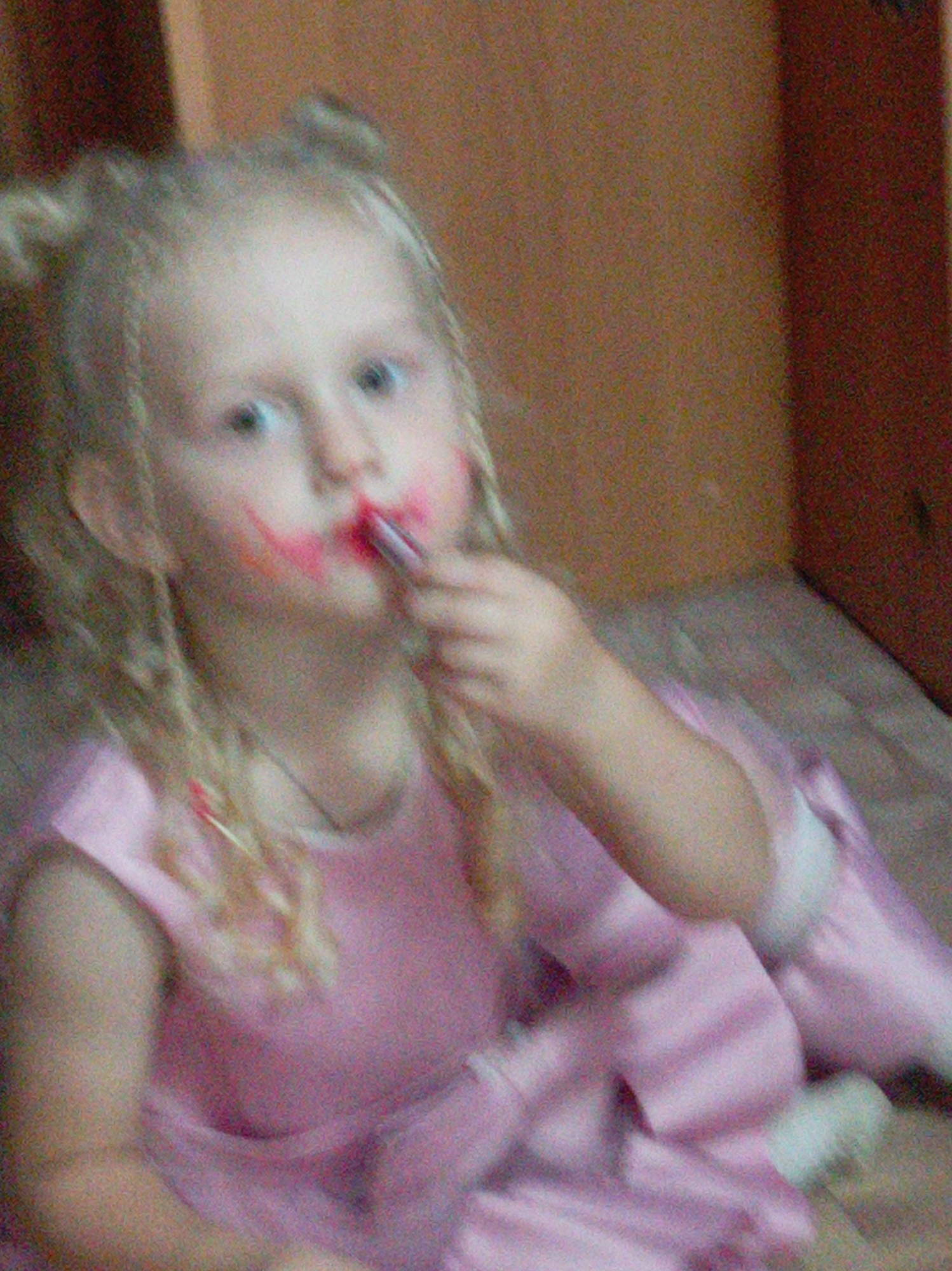 A young girl with blonde hair in pigtails is sitting, wearing a pink dress. She is applying red lipstick to her mouth, with some smudges on her cheeks. Her expression is not clearly discernible due to the low resolution of the image. The background appears to be indoors, with wooden paneling visible. The lighting suggests it might be daytime. The overall impression is of a child playing dress-up.