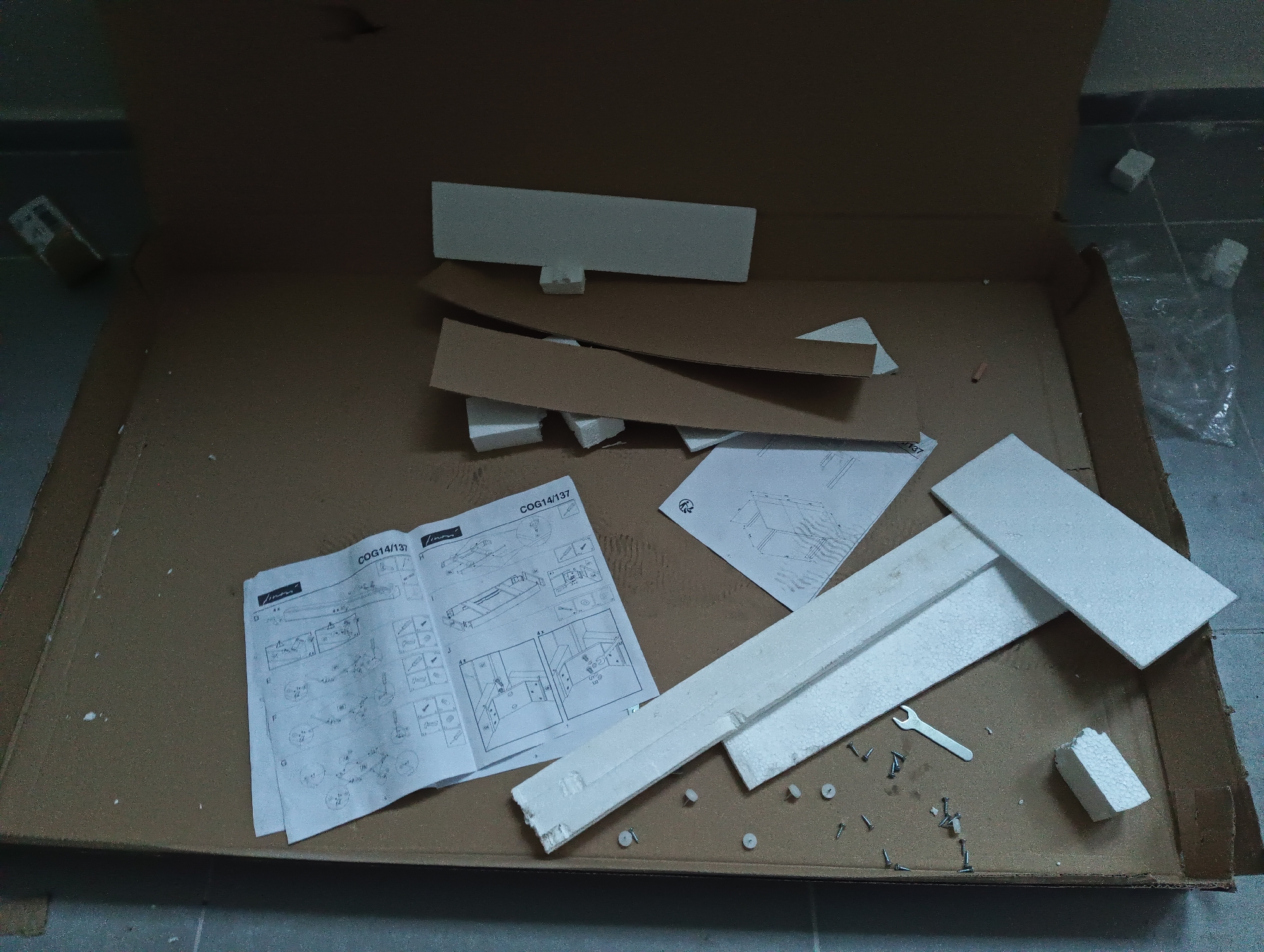 An overhead shot shows the contents of an open brown cardboard box, likely a packaging container for flat-pack furniture, resting on a concrete or tiled floor. The box contains several sheets of assembly instructions, various pieces of white styrofoam packaging, and a collection of small hardware components.
At the center-left, two folded sheets of instruction manuals are prominently displayed. One manual shows diagrams of furniture parts and assembly steps, with the text "COG14/137" and a partially visible brand name "Jimani" at the top. Another smaller sheet to its right also contains diagrams and a circular logo.
Scattered across the bottom-right corner of the box are numerous small screws of different types and sizes, along with several small, white, circular plastic caps or plugs. A small silver-colored metal wrench, typical for assembling flat-pack items, lies among these hardware pieces. Several irregular pieces of broken or intact styrofoam are mixed with the hardware and along the edges of the box, some serving as protective padding.
Other packaging materials, such as curved cardboard pieces, are also inside the box. On the floor outside the box, particularly on the right side, a transparent plastic bag is visible along with more small styrofoam blocks, indicating an active unpacking or assembly process. The overall lighting appears dim, suggesting an indoor setting.