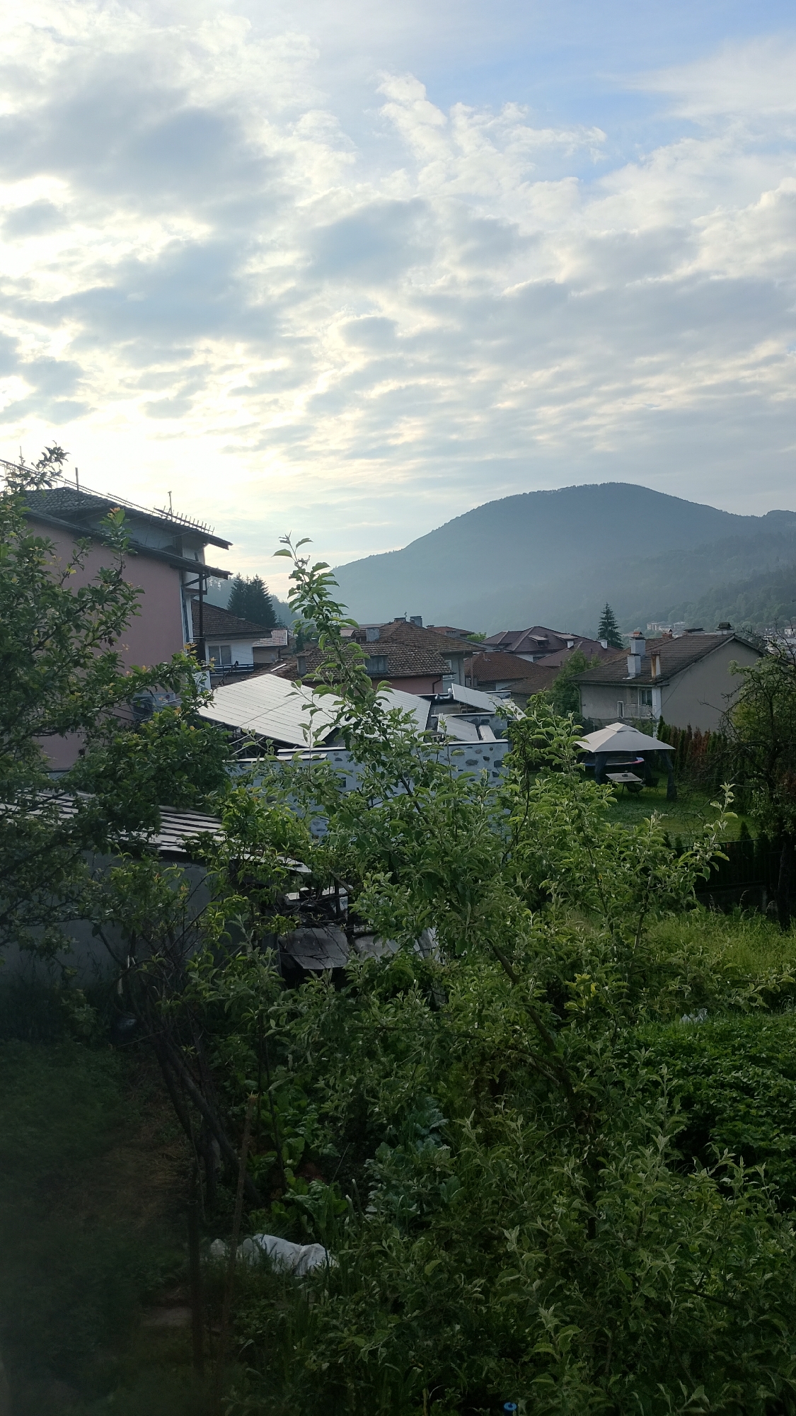 The image depicts a view from an elevated position, looking out over a residential area with a backdrop of rolling hills and a hazy sky. In the foreground, lush green foliage, likely trees and bushes, partially obscures the view.
The middle ground reveals a cluster of houses with terracotta-tiled roofs, characteristic of many European towns. One prominent building in the left foreground has a pink facade and a dark-tiled roof. Adjacent to it, a structure with what appears to be solar panels is visible. Further into the scene, other houses with varying architectural styles can be observed, along with a white gazebo providing shade over a seating area in a garden.
The background is dominated by a large, gently sloping hill, covered in vegetation and fading into a soft haze. The sky above is a pale blue, interspersed with scattered white clouds. The overall impression is of a tranquil, possibly early morning or late afternoon scene in a semi-rural or suburban setting within the city of Smolyan, Bulgaria. The lighting suggests soft, diffused daylight. There are no people or animals visible in the frame.