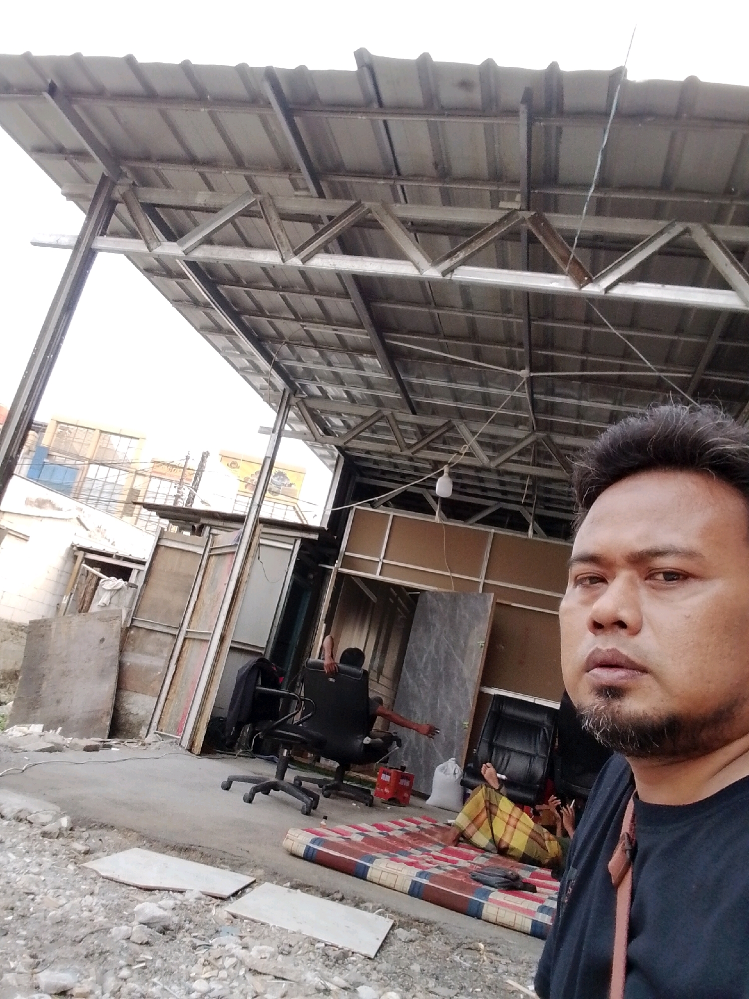 The image captures a scene under a corrugated metal roof supported by a metal truss system. In the foreground, a man is partially visible, facing the camera, with his head and upper torso in focus. He has short, dark hair and a goatee. To his right, a patterned mattress is laid out on the ground. Lying on the mattress is another person, dressed in a yellow and checkered sarong, with their legs extended and smoking a cigarette. In the background, various items are present, including an office chair, what appears to be a stack of wooden boards, and a white sack. Further in the background, buildings with multiple windows are visible, suggesting an urban or semi-urban environment. The overall setting appears to be an open-air workspace or storage area, possibly in Babelan, Indonesia, given the context.