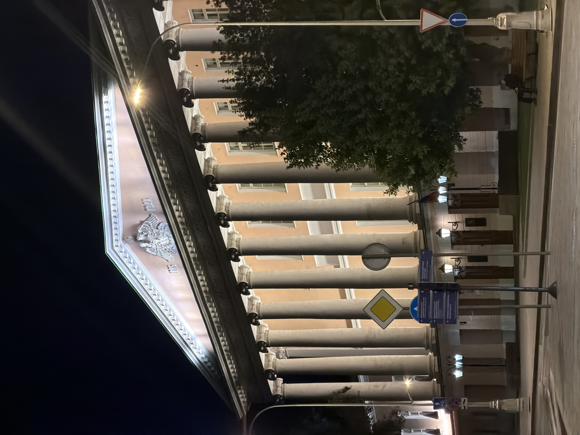 The image shows a neoclassical building at night, identified as the Moscow State University of Culture and Arts (MSUC&A). The building features a prominent portico with a row of tall, fluted Corinthian columns supporting a triangular pediment. Above the columns, the pediment is adorned with a large, ornate eagle crest and the inscription "1775 ГОДА" (Year 1775). The facade of the building is illuminated, highlighting its architectural details.
In the foreground, there is a street with a sidewalk. Several street signs are visible, including a diamond-shaped yellow sign, a blue directional signpost with text in Russian, and a triangular yield sign with a blue circular arrow. Lampposts with multiple lights are also present, casting a warm glow on the scene. A few trees, one large and dense, stand to the right of the building, partially obscuring the lower floors.
A single person, dressed in dark clothing, is seated on a park bench on the sidewalk, facing away from the camera and towards the building. The scene is characteristic of an urban setting at night, with the building being a significant architectural landmark in Moscow.