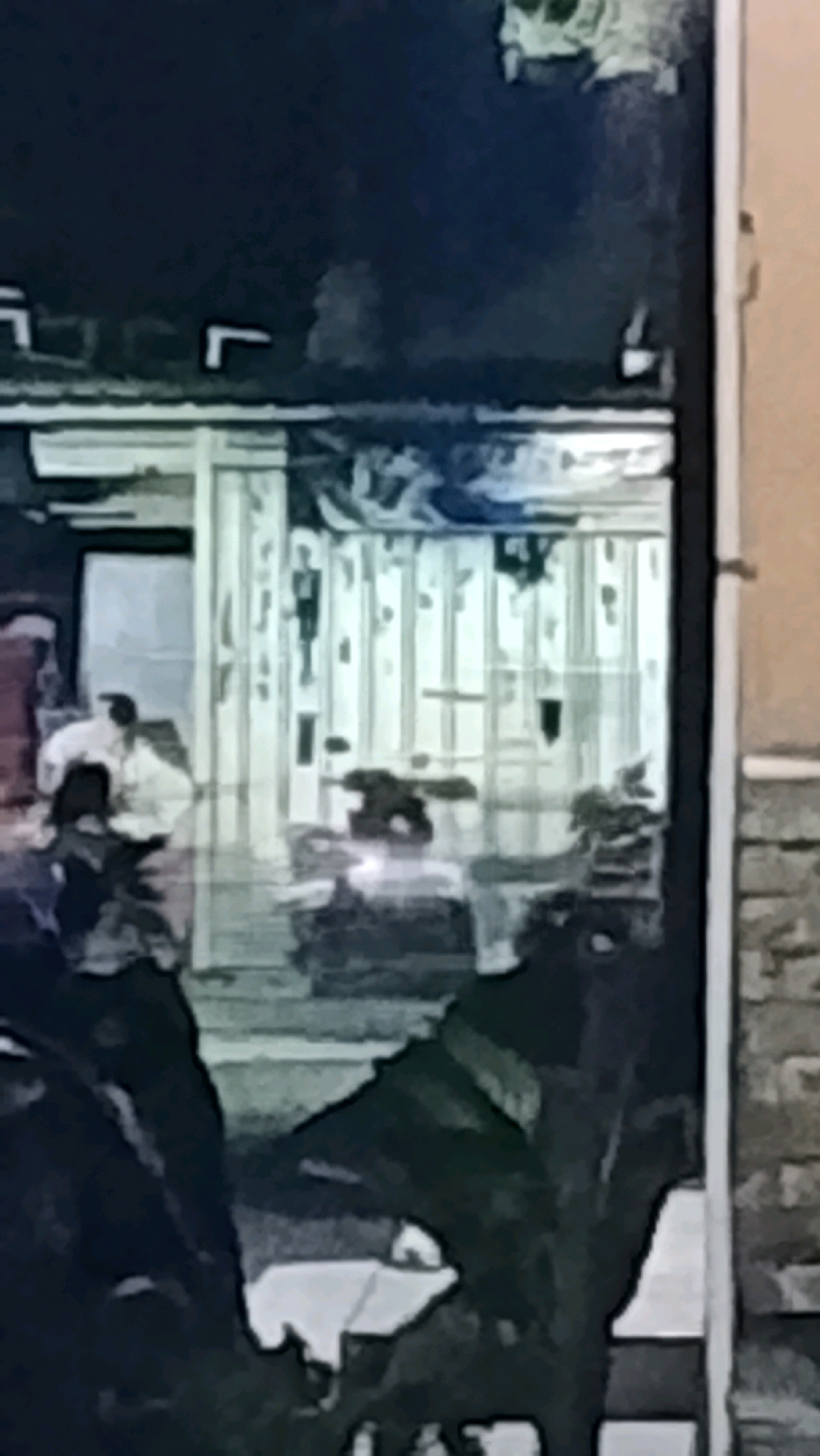 The image captures a nighttime scene through what appears to be a glass-fronted establishment, possibly a shop or restaurant, located in Ujungrusi, Indonesia. The exterior shows a dark, possibly tiled or brick wall on the right. The interior is dimly lit, revealing a row of vertical, light-colored panels or blinds creating a striped effect.
Inside, several figures are visible. In the foreground, to the left, a person is seated with their back to the viewer, wearing a light-colored shirt and possibly pants. To their right, a dark, shadowy object appears to be a table with something on it, possibly a laptop or other electronic device, emitting a bright light. Further back and to the right, another figure, also in light-colored clothing, is seated. There are dark shapes of plants or furniture in the immediate foreground, obscuring the lower part of the scene. The overall impression is a blurry, low-light photograph of an indoor scene at night.