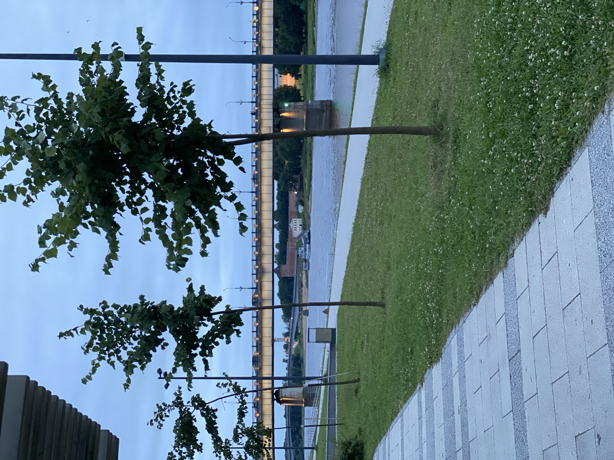 The image captures a scene along the waterfront in Veliky Novgorod, Russia, likely during twilight. In the foreground, a modern pedestrian walkway with light-colored paving stones runs diagonally from the lower left corner upwards. Adjacent to the walkway is a neatly mowed grassy area dotted with small white flowers. Several young, slender trees with green leaves are planted in the grass.
Dominating the middle ground is a wide, calm river with gentle ripples on its surface. Across the river, a long, multi-span bridge is illuminated with lights, suggesting traffic movement. The bridge is supported by robust concrete pillars. On the far bank of the river, there are various buildings, including what appears to be a historical fortress or castle with brick towers. Trees line the riverbank on the opposite side.
In the background, the sky is a muted, overcast blue-grey, indicating either early morning or late evening. There are no specific individuals prominently featured in the image, though a distant figure in an orange garment can be observed sitting on the grass near the riverbank. The overall atmosphere is serene and urban, highlighting a blend of modern infrastructure and natural elements against a historical backdrop.