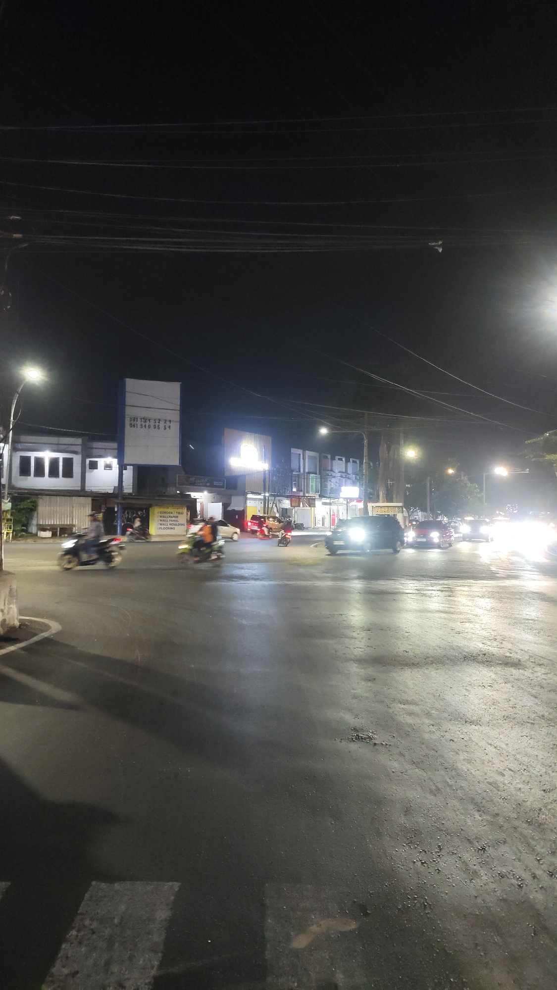 This is a nighttime, eye-level shot of a street intersection in Malang, Indonesia. The scene is illuminated by streetlights and the headlights of vehicles. Several motorbikes and cars are visible, some in motion, creating motion blur. The road surface appears wet, reflecting the lights. Buildings line one side of the intersection, displaying various signs and illuminated windows. Overhead, power lines crisscross the dark sky. In the foreground, a pedestrian crossing is marked on the asphalt.