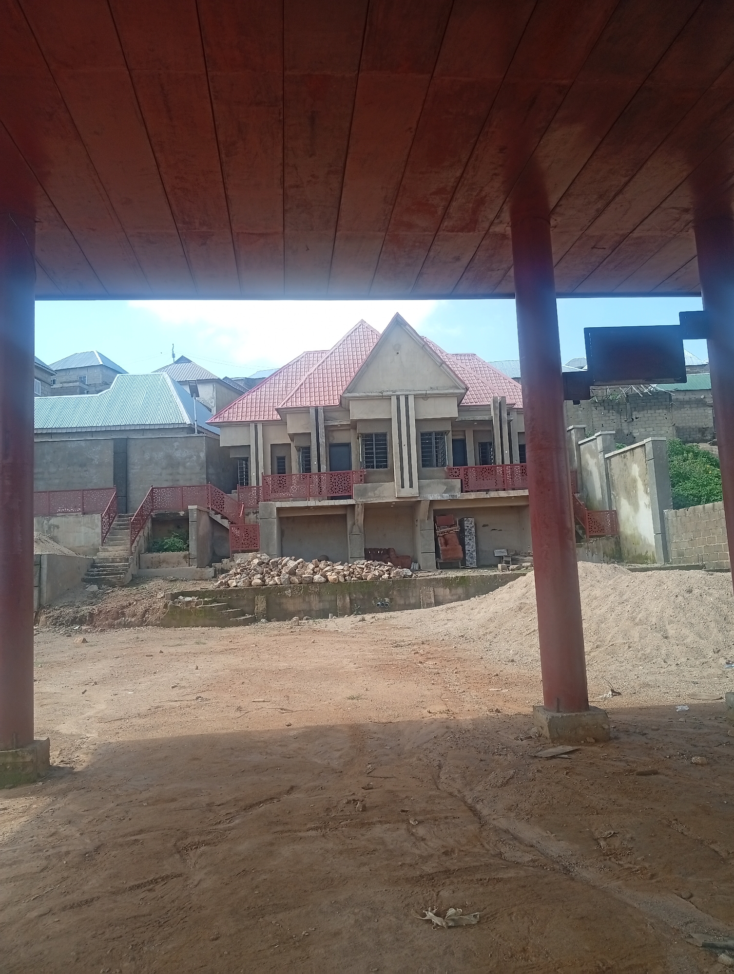 The image shows a partially constructed building with a red tiled roof and beige-colored exterior walls. The building has multiple windows and balconies with decorative red railings. In front of the building, there is a pile of rocks and construction materials. To the left of the main building, another building with a silver corrugated metal roof is visible. The scene appears to be an outdoor construction site with a dirt ground in the foreground. The sky is visible in the background with some clouds. The vantage point of the image is from beneath a wooden overhang, supported by thick red-colored columns. This specific scene is located in Jos, Nigeria.