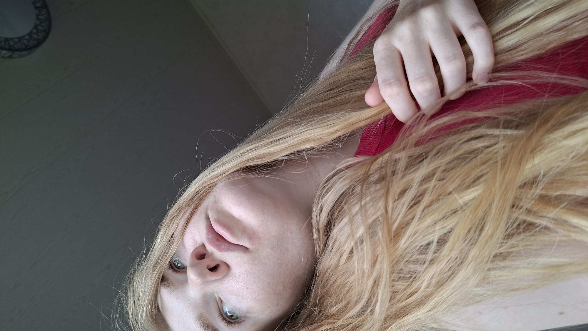 An angled, close-up indoor portrait features a person with long, blonde hair and light-colored eyes. The image is significantly tilted, giving the impression that the person is looking upwards or lying down while facing the camera. The subject's face occupies the lower right portion of the frame, with the left side of the face and upper body mostly obscured by hair or cropped out.
The person has fair skin, visible freckles on the nose and cheeks, and distinct green or hazel eyes that are open and looking towards the viewer. The lips are slightly parted, revealing no teeth. Long strands of blonde hair, appearing to be light golden or strawberry blonde, flow across the frame, covering much of the person's neck and shoulder area. A hand, with visible fingers, gently holds or separates a portion of the hair near the top right of the image, revealing a vibrant red fabric underneath, possibly part of clothing or an accessory.
The background consists of a dark, muted gray surface, likely a ceiling or wall, showing faint lines that could indicate paneling or texture. In the upper left corner, a partial view of a metallic, geometric light fixture or ceiling fan is visible. The lighting is soft and even, suggesting either natural light from an unseen window or diffused artificial lighting. No text is visible in the image. The overall mood is calm and contemplative.