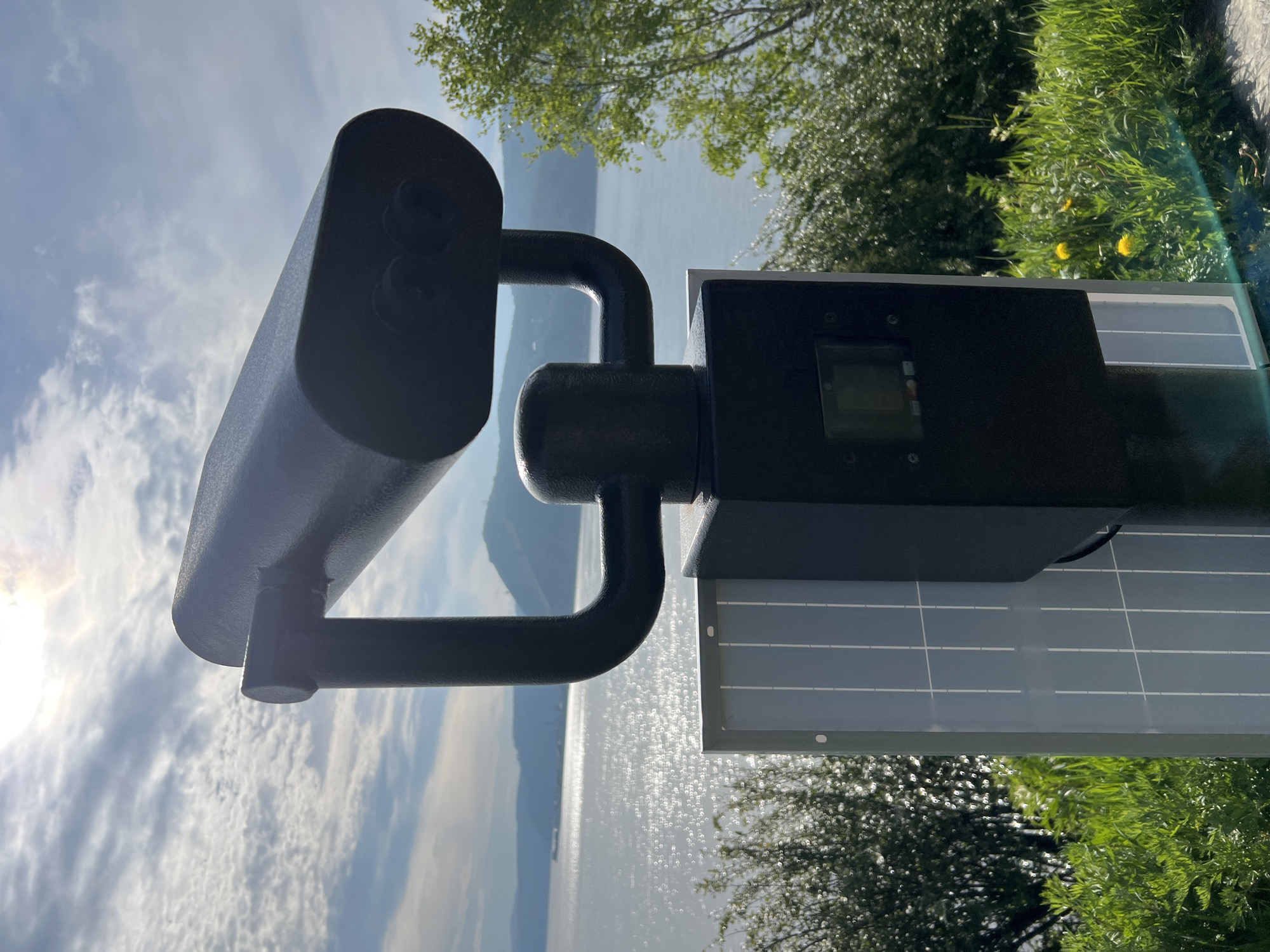 A dark, heavy-duty binocular viewer stands on a black pedestal in the foreground, angled slightly to the right. The viewer is powered by a solar panel attached to the front of the pedestal. The background features a wide, sun-drenched body of water, likely Lake Baikal given the location in Listvyanka, Russia, with distant hills and a hazy sky with scattered clouds. Green foliage, including grass and trees, surrounds the viewer, with a few yellow dandelions visible in the lower right corner. The scene appears to be outdoors, in a scenic overlook.