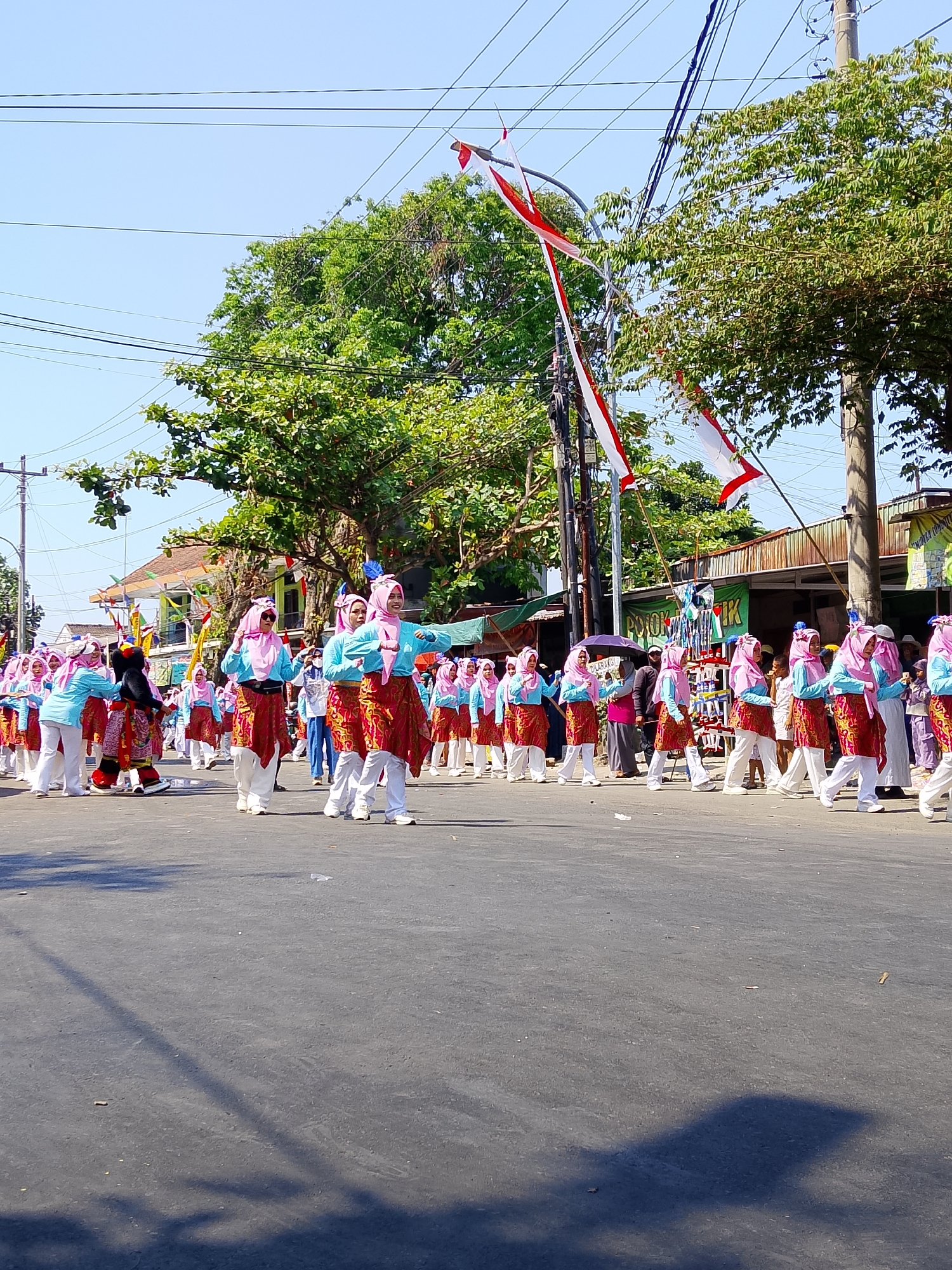 In Balun, Indonesia, a parade is underway on a paved road under a clear blue sky. A group of individuals, primarily young women wearing pink headscarves and light blue tops with red and gold patterned skirts, are marching in formation. They are accompanied by one person in a black mascot costume with yellow accents. Red and white flags, likely representing the Indonesian flag, are displayed on poles along the parade route. Trees line the sides of the road, and buildings with colorful decorations are visible in the background. The event appears to be a community celebration or festival.