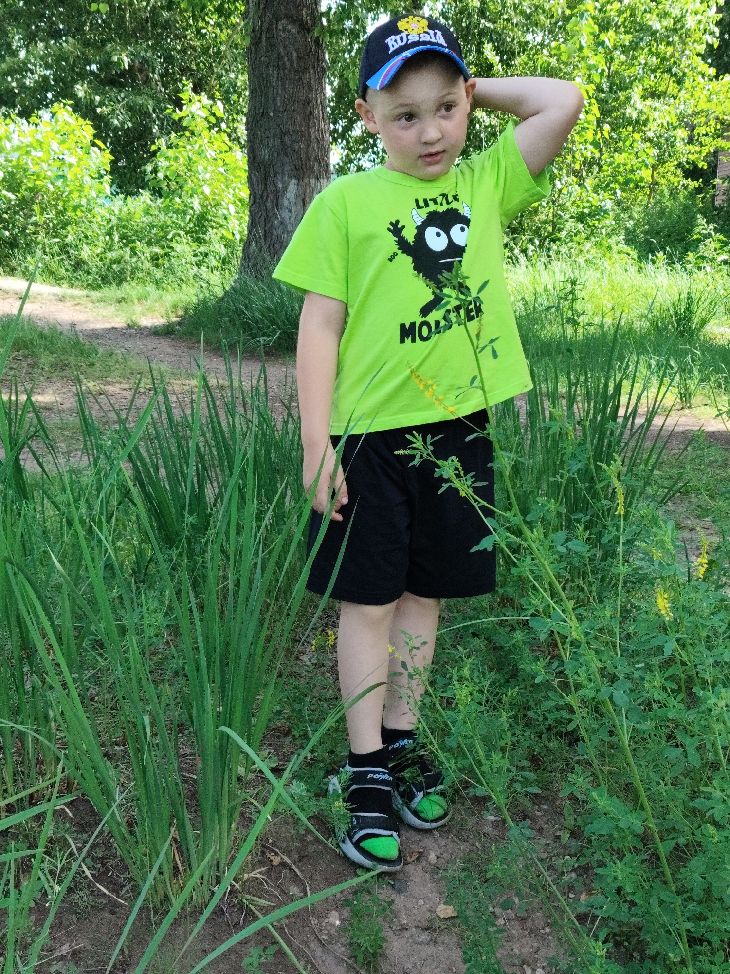 In the city of Raikov, Russia, a young boy stands amidst lush greenery. He is wearing a bright neon green t-shirt with a cartoon monster graphic and the word "MONSTER" printed below it, paired with black shorts. A dark baseball cap with "RUSSIA" written on it adorns his head. His right arm is raised, touching the back of his head. He wears black socks with "POWER" written on them and black and green sneakers. The environment is a natural setting, likely a park or wooded area, with tall grass and trees in the background. The ground appears to be a dirt path.