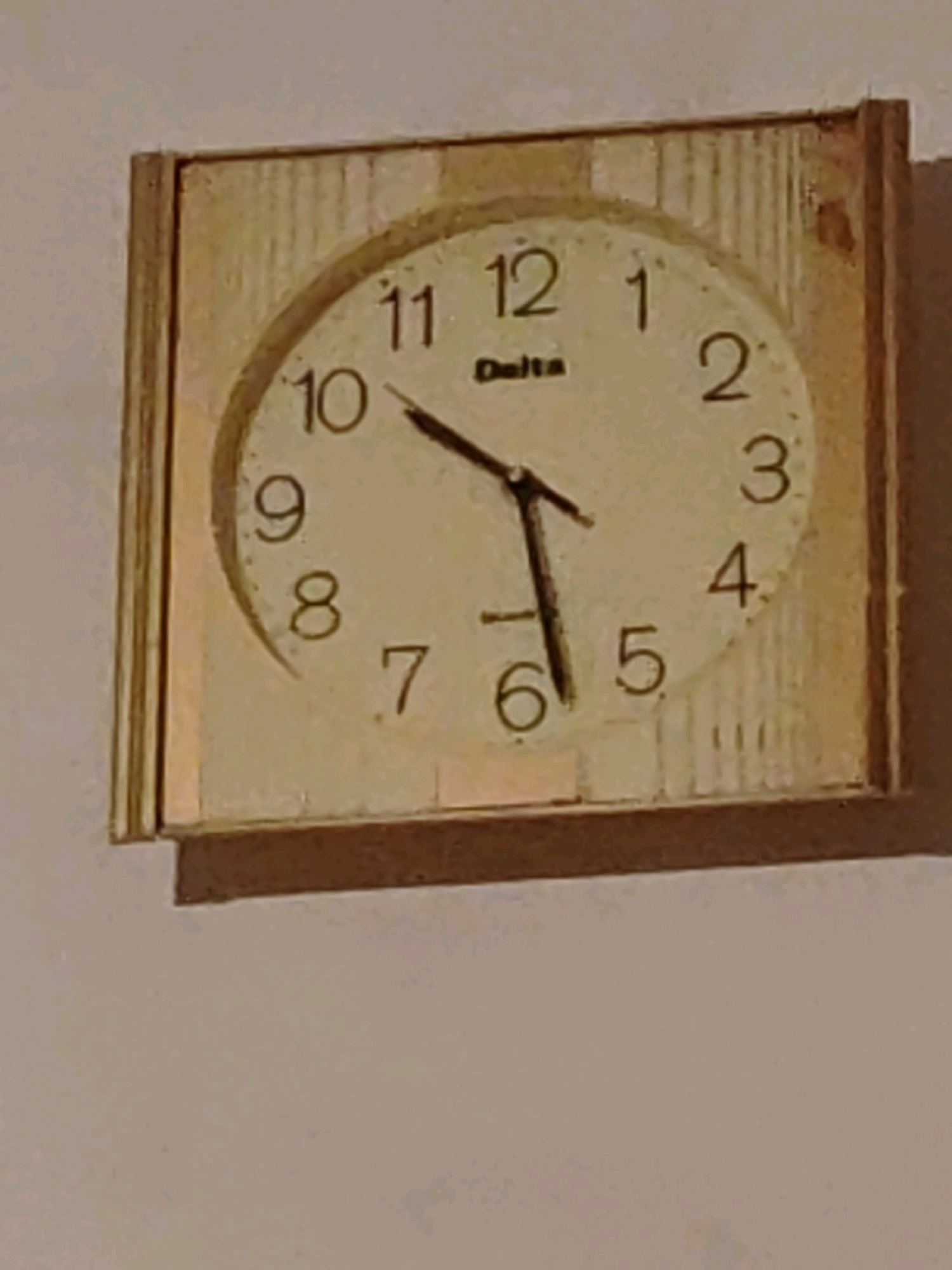 A close-up, slightly blurry image shows a wall clock with a light-colored face and a wooden frame. The clock has black Arabic numerals indicating the hours, and black hands. The hour hand points to approximately 10, and the minute hand points to approximately 57. The brand name "Delta" is visible just below the 12. The clock is mounted on a plain, light-colored wall. The bottom of the image shows a dark brown ledge or shelf attached to the wall, possibly a windowsill. No people are visible in the image. The image quality is not sufficient to determine the exact location within Ninotsminda, Georgia.