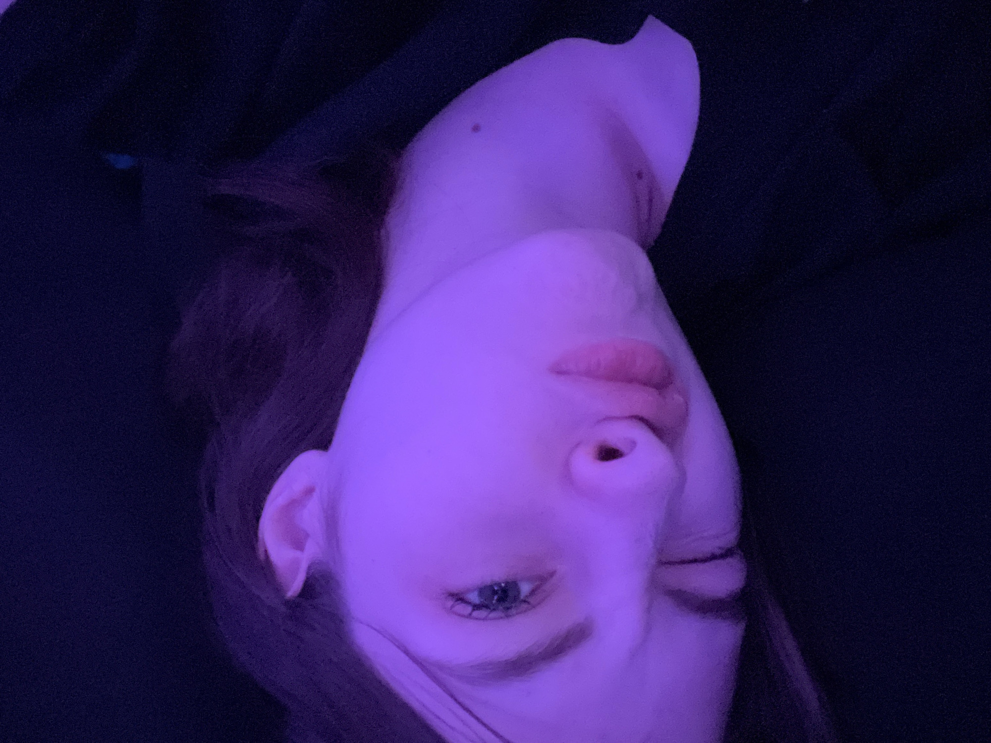 The image shows a person's face, illuminated with a purple light. The person appears to be lying down, as the head is oriented sideways. One eye is open and visible, while the other is closed or partially obscured. The person's lips are visible, and a mole is present on their neck. The background is dark and indistinct. There is no location information in the photo.