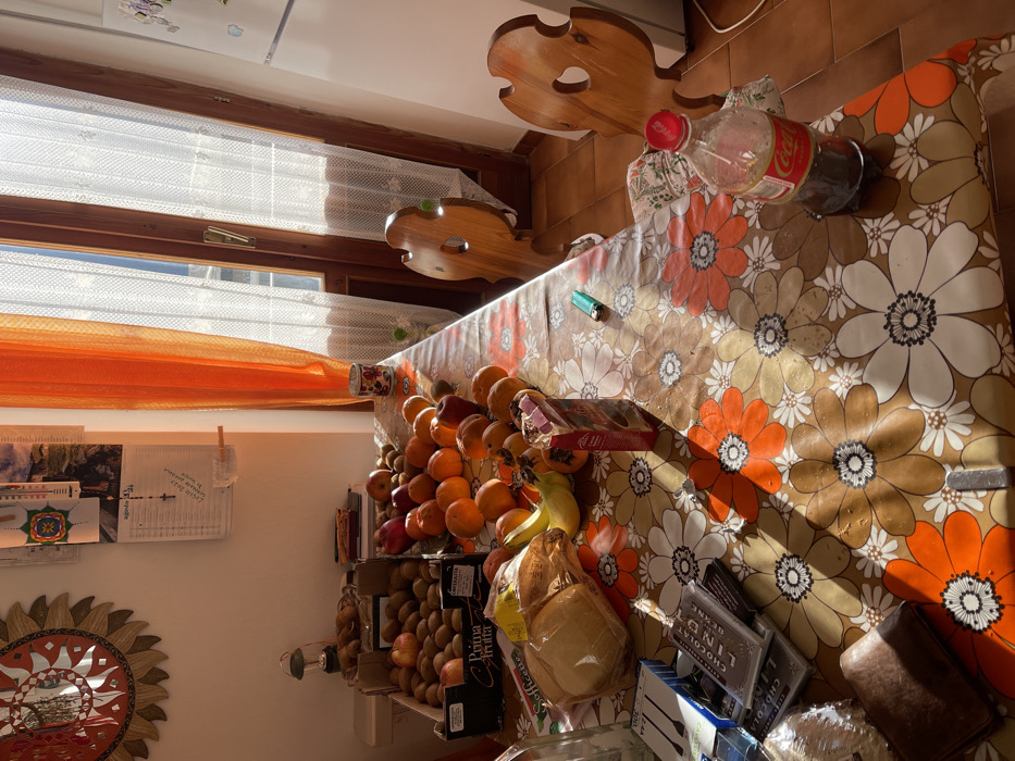 The image, captured in a rotated landscape orientation, depicts a sunlit domestic interior, likely a kitchen or dining area, in Torre di Santa Maria, Italy. A large wooden table, adorned with a vibrant floral tablecloth featuring orange, white, and brown blossoms, dominates the scene.

On the table, a substantial display of fresh fruit includes a large pile of oranges, several red apples, and a bunch of yellow bananas. Below the table, boxes containing kiwis, some labeled "Prima Frutta," are visible. Other food items present are two plastic bags of bread (one labeled "Coffeette"), multiple boxes of "Lindt" chocolate, and a smaller box of confectionery. A partially consumed Coca-Cola bottle with a red cap and a green disposable lighter also rest on the tablecloth. A white ceramic mug is placed near the window.

Bright, direct sunlight streams through a large, wooden-framed window, illuminating the scene and casting sharp shadows across the table. The window is dressed with white sheer curtains and an orange sheer fabric drape, adding a warm glow. Above the table, decorative carved wooden elements are affixed to the wall. On a lighter-colored wall to the left, a prominent, circular sun-shaped mirror is mounted. Papers, including one with partially visible Italian text like "LUNEDÌ 2023," are pinned to the wall.

No people are present, suggesting a quiet moment in a well-stocked home. The bright lighting indicates it is daytime, possibly morning or afternoon. The overall impression is one of warmth and abundance in a traditional setting.