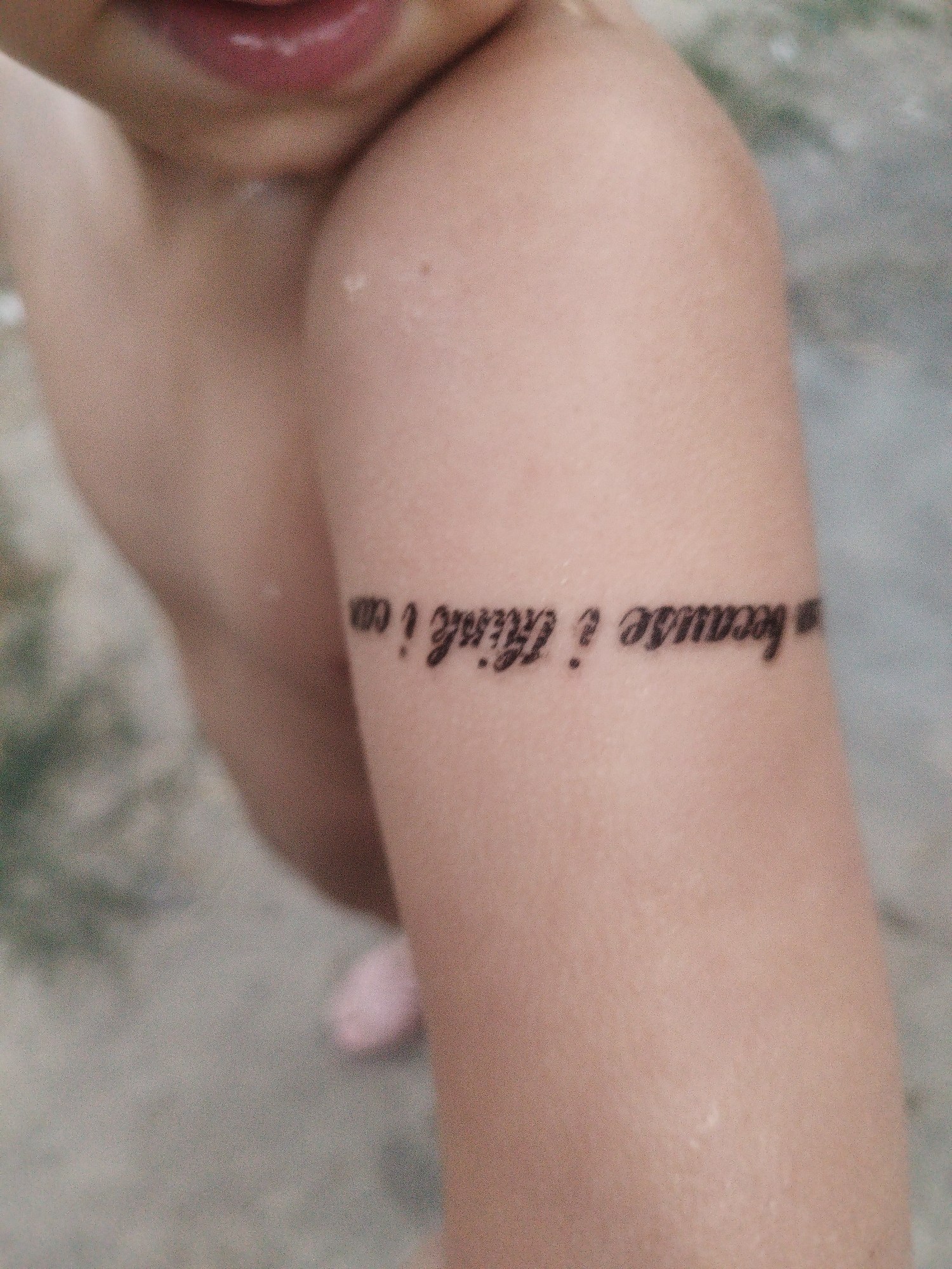 The image shows a close-up of a person's arm with a tattoo. The tattoo is a phrase written in cursive script and appears to say "because i think i can". The arm is lightly tanned and shows some texture. In the background, there is a blurry greyish surface that could be pavement or ground. The person's lips and chin are partially visible at the top of the frame. There is no indication of the location being in the city of Shymkent, Kazakhstan.