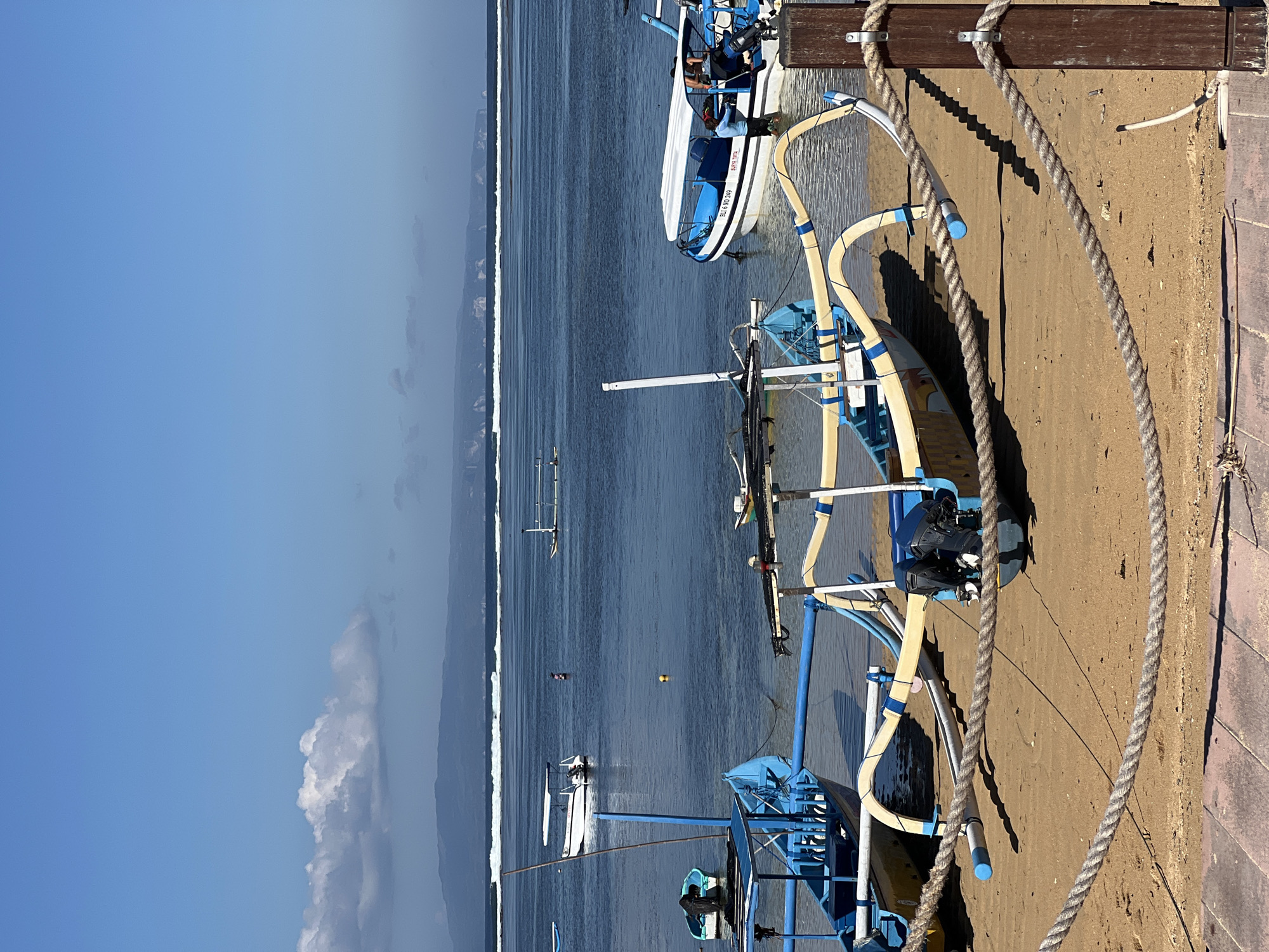 The image shows a tropical beach scene in Denpasar, Indonesia, likely Sanur Beach, on a clear day with a bright blue sky and scattered clouds. Several traditional outrigger boats, known as "Jukung," are pulled up onto the sandy shore. These boats are characterized by their distinctive bamboo outriggers painted yellow and blue. In the foreground, a thick rope barrier is visible, strung between wooden posts.
In the water, more Jukung boats are anchored or floating. One larger boat with a white canopy and blue hull, identified by the text "BLI 6 NO 249" and "SURYA TIRTA" on its side, has several people on board, appearing to be passengers or crew preparing for or disembarking from a journey. The water is calm with gentle waves breaking in the distance.
Further out at sea, a hazy mountainous coastline is visible on the horizon. The overall impression is one of a serene coastal environment, a common sight at tourist beaches in Denpasar, where these traditional fishing and transport vessels are prevalent. The lighting suggests it is daytime, with sunlight casting shadows on the sand.
