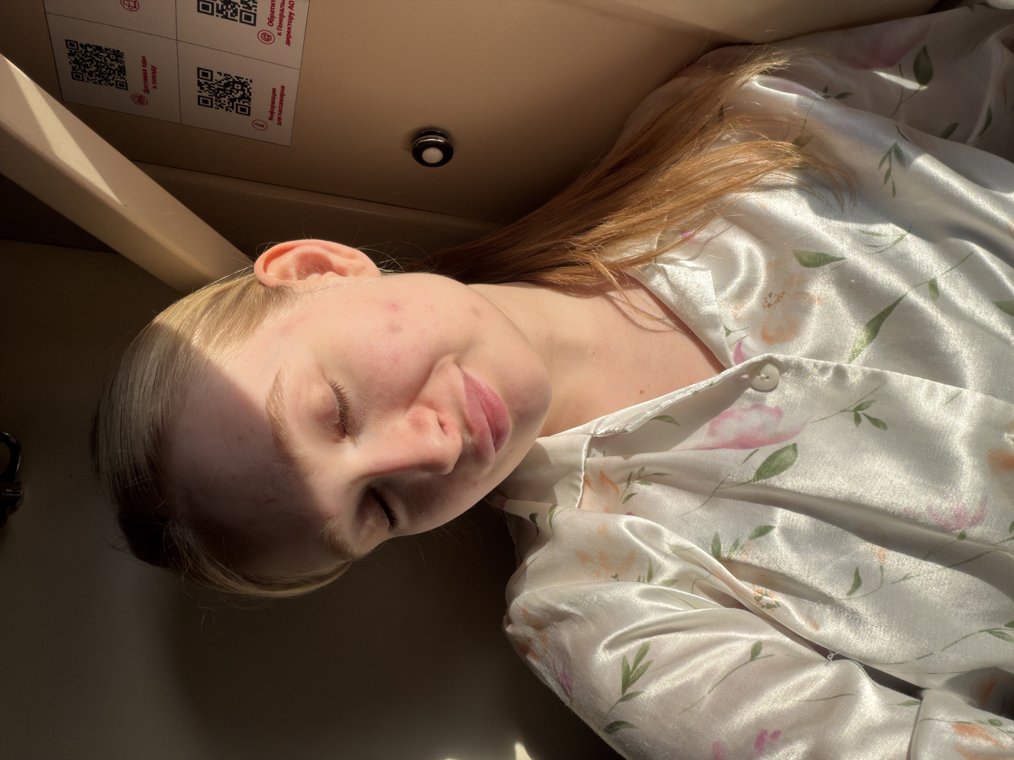 A fair-skinned young person lies in an enclosed space, possibly a compartment. The person has blonde hair with reddish-brown streaks, closed eyes, and slightly parted lips. The person's skin has some redness and small blemishes. They are wearing a light-colored satin-like garment with a floral pattern. The background includes parts of a ceiling with a small light fixture and labels with QR codes. The image was taken in a compartment in Krasnyy Sulin, Russia.