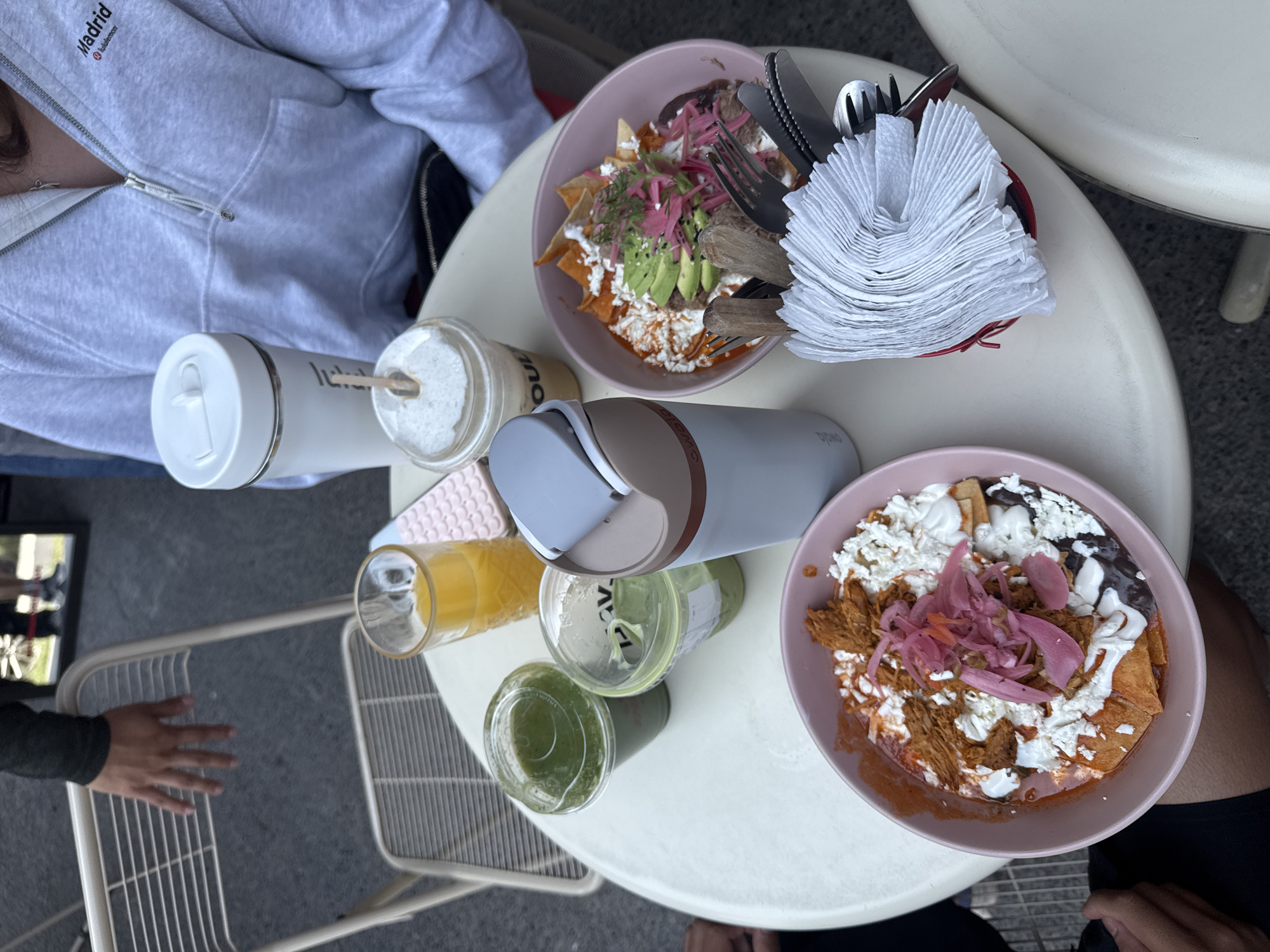 An overhead view captures an outdoor dining scene at a cafe in Puebla, Mexico, featuring food and drinks on a light-colored round table. Two individuals are partially visible: one wearing a light grey zip-up hoodie with "Madrid lululemon" text on the chest, and another whose leg and arm are seen.
On the table are two pink bowls filled with what appear to be chilaquiles. The top bowl contains tortilla chips covered in a red sauce, topped with crumbled white cheese, sliced avocado, and pickled red onions. The bottom-right bowl also features tortilla chips with red sauce, white cheese, black beans, shredded meat (possibly tinga), and pickled red onions.
A variety of beverages are present: a white thermos, a glass with a frothy light-colored drink (bearing the partial text "SOUL") and a straw, a grey and beige thermos with "DiGNO" visible, a glass of orange juice, and two clear cups containing a green liquid, likely a juice or smoothie. A red holder with metal cutlery and a substantial stack of white napkins sits next to one of the food bowls. The scene is brightly lit, indicating daytime, and light-colored metal chairs are visible around the table on a paved surface.