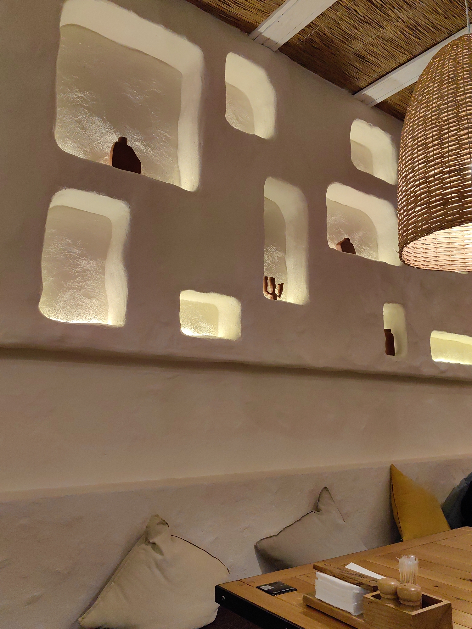 The image depicts an indoor dining establishment in Sochi, Russia, characterized by its Mediterranean-inspired aesthetic. No individuals are present.
The focal point is a textured, off-white wall featuring multiple irregularly shaped, recessed niches. These niches are illuminated from within by hidden light sources, casting a soft, warm glow. Several niches contain dark, silhouette-like decorative objects; these include a squat, rounded form in the upper left, an abstract, hand-like sculpture in the middle left, a narrow bottle-shaped object in the middle right, and a small vase-like item in the lower right.
Above, the ceiling consists of exposed white wooden beams against a background of woven straw or bamboo matting. A large, bell-shaped pendant lamp, crafted from woven wicker or rattan, hangs prominently on the right side of the frame, emitting a warm light downwards.
Below the wall decor, a long, built-in bench seat, matching the wall's texture and color, is furnished with several cushions. These include multiple off-white or cream-colored cushions, a single bright yellow cushion, and a dark blue or grey cushion towards the far right.
In the foreground, a light-toned wooden table with visible slats occupies the lower right quadrant. On the table, a wooden caddy holds a stack of white paper napkins, two small wooden shakers (likely salt and pepper), and a container filled with white toothpicks. To the left of the caddy, a black rectangular card, possibly a menu or QR code, is placed on the table.
