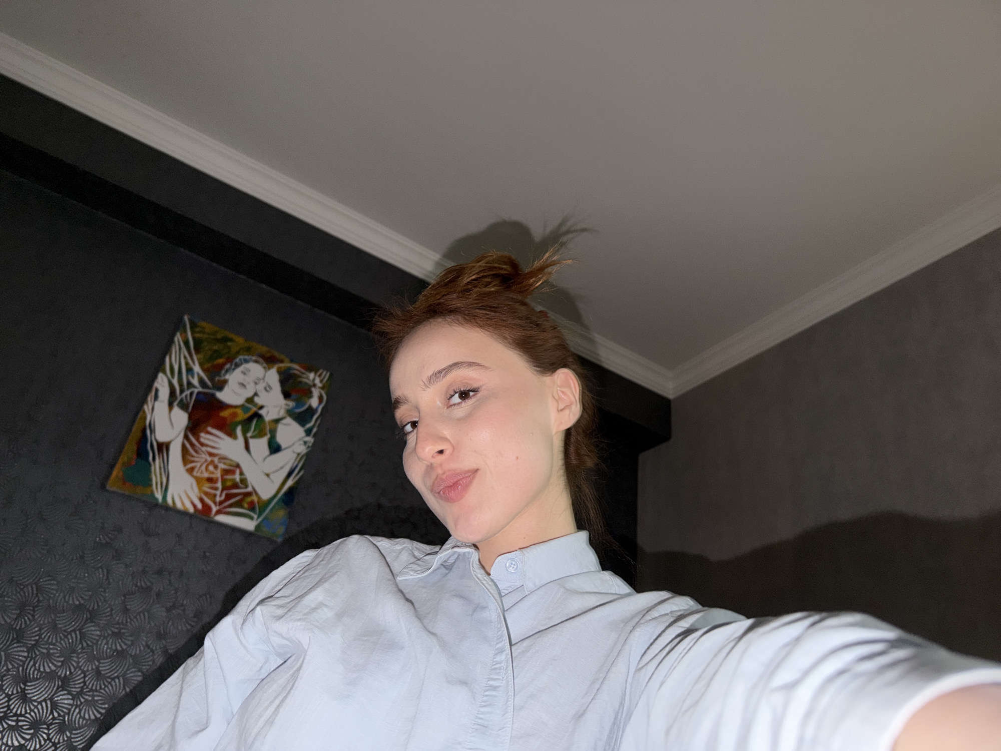 A selfie taken from a low angle captures a woman with reddish-brown hair pulled into a messy bun. She wears a light blue button-up shirt. The background features a dark headboard and gray patterned wallpaper. On the wall, there is a colorful painting depicting two figures embracing. The scene is located indoors. The city of Tbilisi, Georgia, cannot be determined from the image.