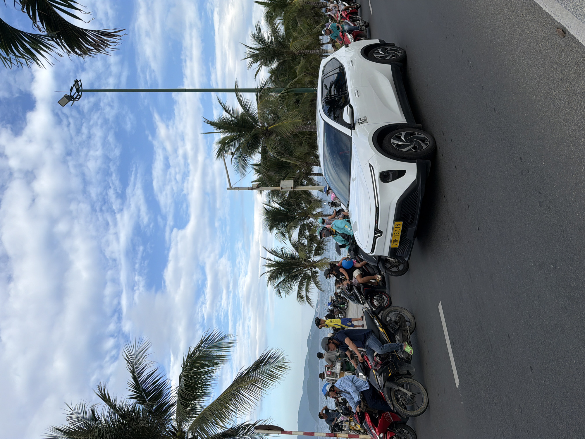 In Nha Trang, Vietnam, a white electric vehicle drives down a coastal road lined with palm trees under a cloudy blue sky. The vehicle, a VF8 electric SUV, is in the foreground on the right, with its license plate reading "79H-137.15". A group of motorcycles and their riders are in motion on the road, with some vehicles and people visible on the sidewalk and beyond, near the sea. The atmosphere is bright, suggesting daytime, and the scene captures a typical street view in a tropical coastal city.