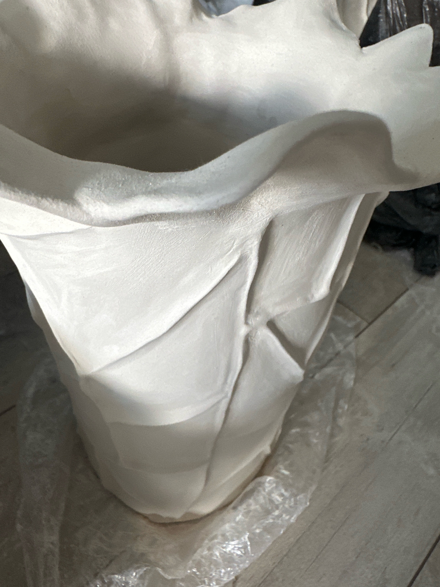 The image shows a close-up of a white, unfinished ceramic vase or pot. The vase has a textured surface with visible seams and folds, giving it a sculpted appearance. It is partially wrapped in clear plastic sheeting, and rests on a wooden floor. In the background, a dark object, possibly a bag, is visible. The lighting suggests an indoor environment. The image does not contain any information that would allow for its location to be specified in Nevinnomyssk, Russia.