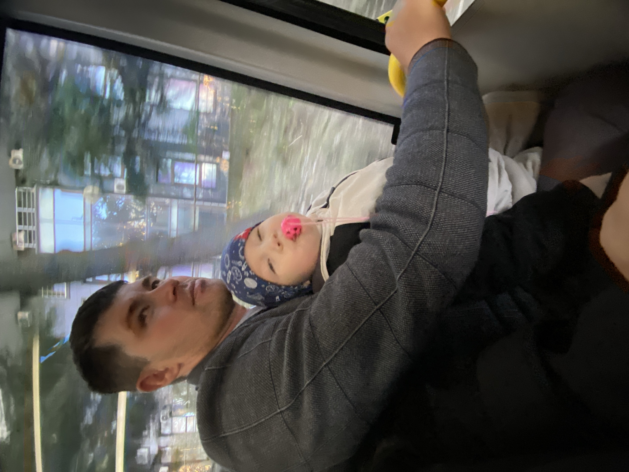 The image shows a man on a bus in Sochi, Russia. He is holding a baby who is wearing a blue bandana and has a pink pacifier in their mouth. The man is wearing a dark jacket and holding onto a yellow handrail. Outside the window, there are buildings and trees reflecting in the window. The man appears to be looking upward.