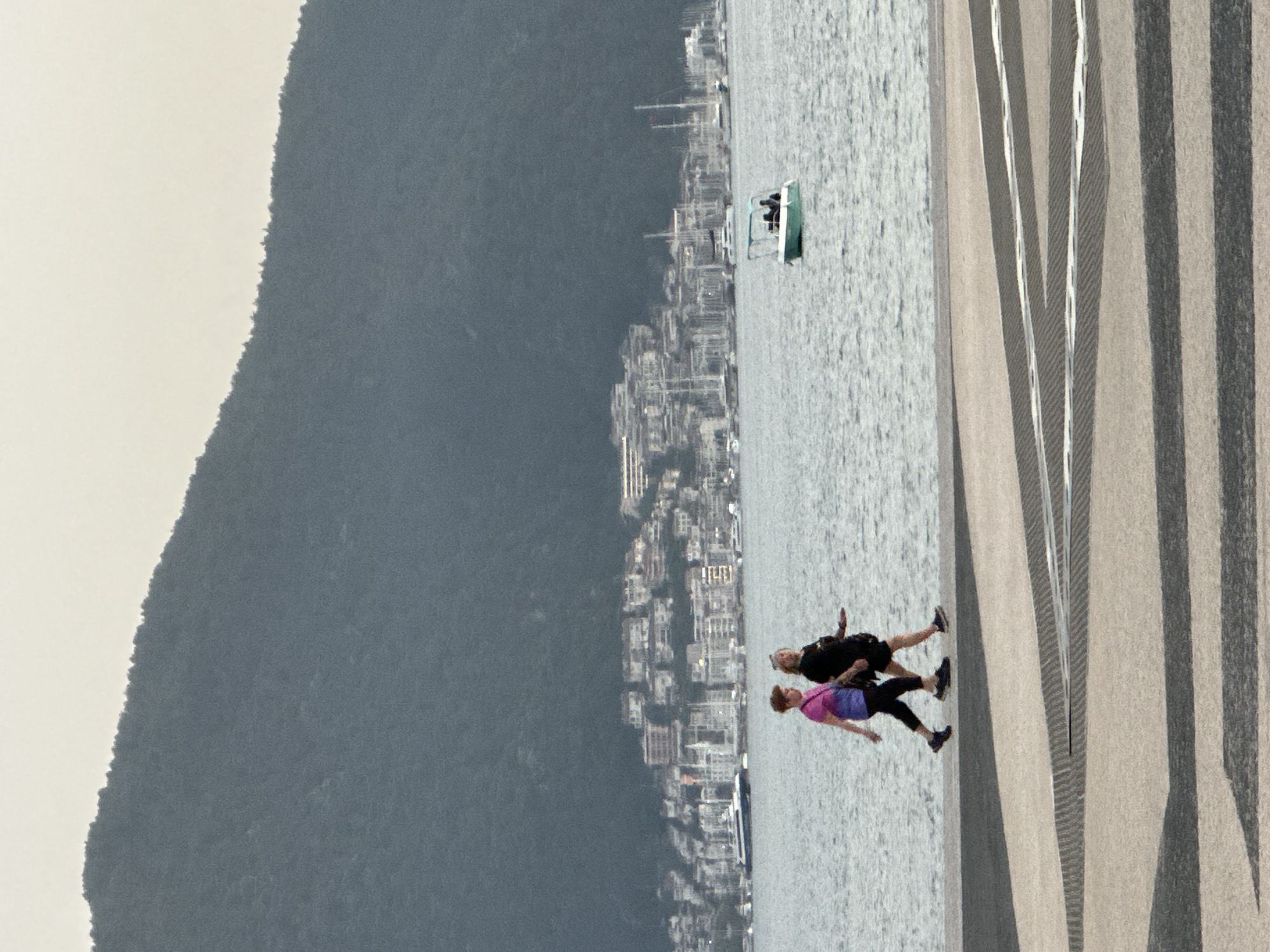 The scene is a high-angle shot of a coastal area, potentially in Fethiye, Turkey. In the background is a dark, mountainous landform, possibly a hill or mountain, adjacent to an urban sprawl. The waters of a bay are prominent.
Two people, an older woman and man, are walking on a promenade. The woman is wearing a pink shirt and the man a black jacket.
A small boat with passengers is present in the water. A grey and white striped promenade extends along the coastline, forming a walkway for pedestrians.