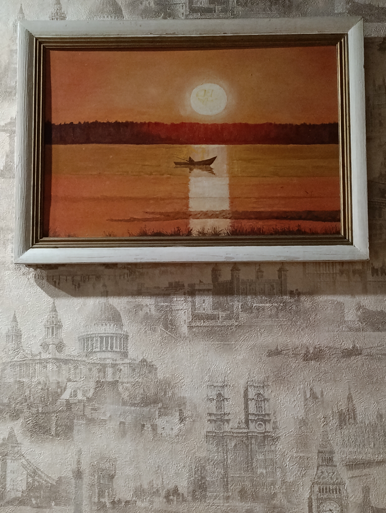 A framed painting hangs on a textured wallpaper featuring a cityscape, likely depicting London. The painting depicts a sunset over water, with a silhouetted boat in the center of the water and the sun reflecting on the water. The overall color scheme is warm, with shades of orange and brown dominating the scene.
It is impossible to know the location in Krasnoyarsk, Russia from this image.