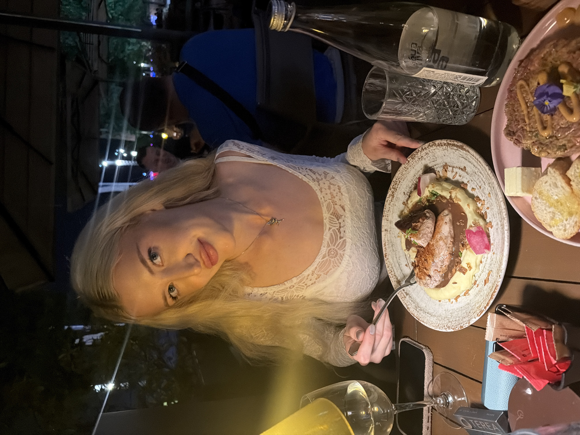 The image shows a blonde woman sitting at a wooden table in a restaurant. She is wearing a white lace top and a silver necklace. On the table are two plates of food, a bottle of water, a glass, a fork, a smartphone, a wine glass, and sugar packets. The plates contain a dish with meat and mashed potatoes and a pink plate containing raw beef tartar with slices of bread. The location may be Belgrade, Serbia. A man is seated in the background.