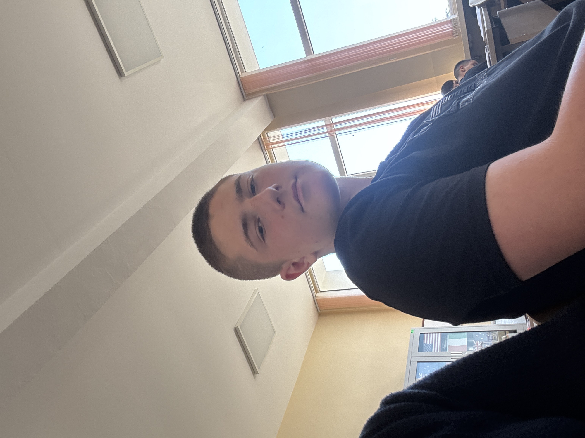 The image shows a young Caucasian male with a buzz cut, wearing a black t-shirt, in what appears to be a classroom. He is looking towards the camera with a neutral expression. His right arm and shoulder are visible in the foreground, and he is seated at a desk. Behind him, to the left, a filing cabinet with flags is partially visible, suggesting it might be a classroom in Stara Zagora, Bulgaria, possibly a school with international affiliations or a classroom displaying international symbols. To the right, a window with sheer curtains allows sunlight to enter, illuminating the room. Other students are visible in the background, seated at desks. The ceiling features fluorescent lighting panels and a sloped design. The overall environment suggests a daytime setting in an educational institution.