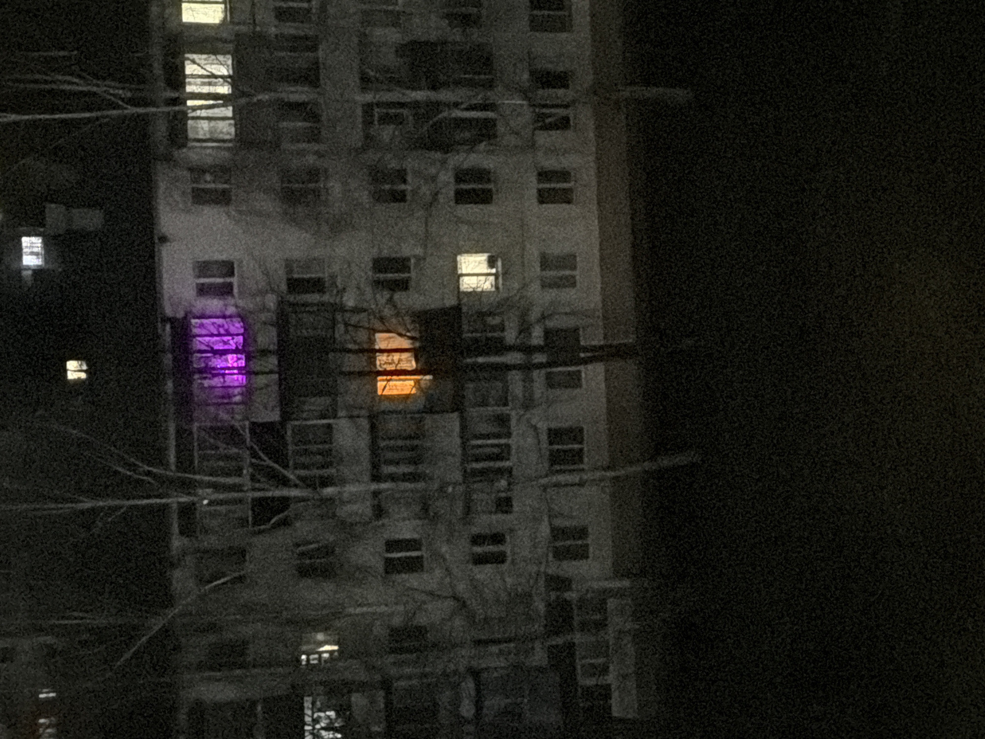 A nighttime image of an apartment building. Two windows on the building are lit up. One window shows a purple light and another shows an orange light. There are tree branches in front of the building. The building is likely located in the city of Chaikovsky, Russia.
