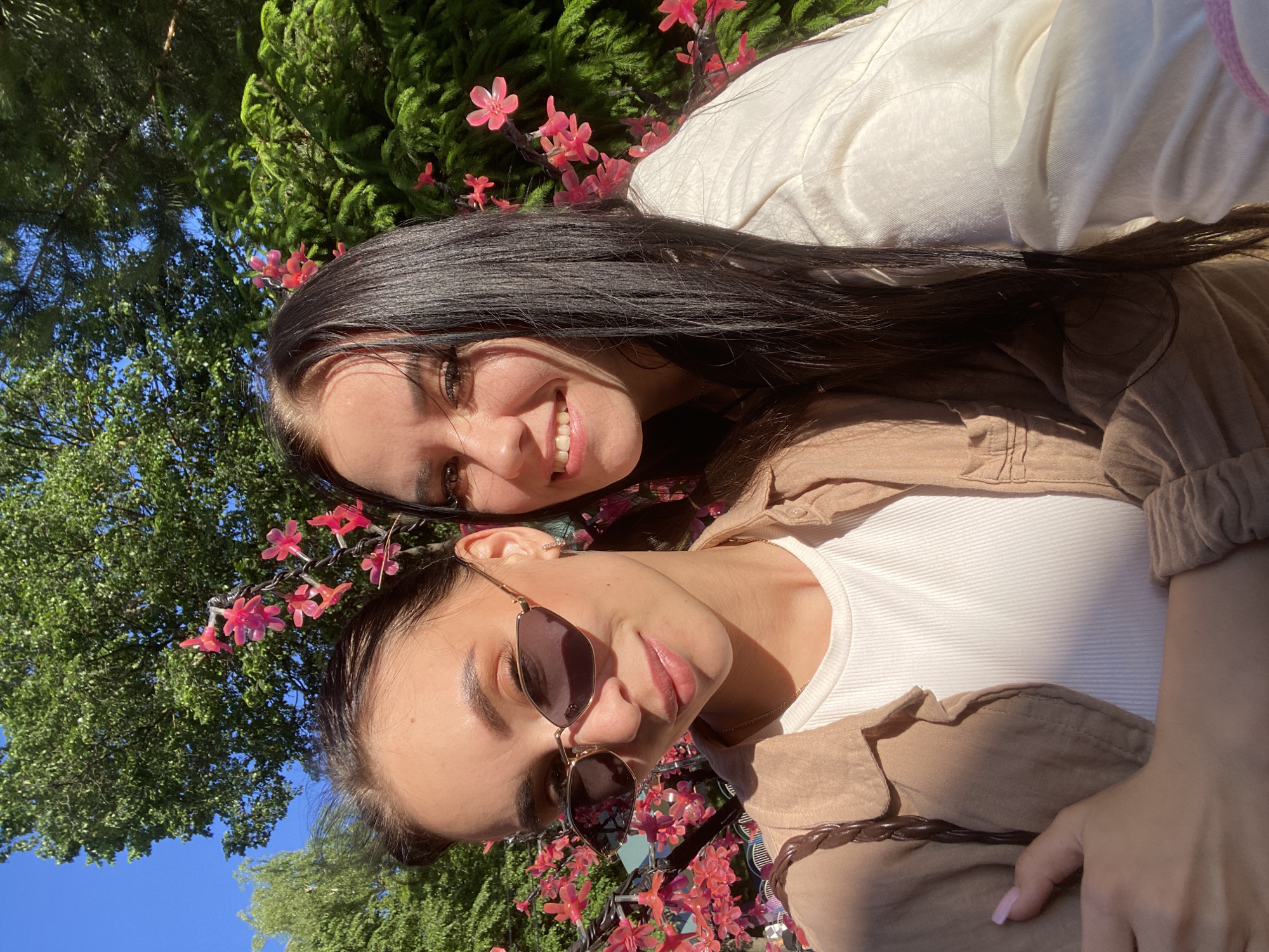 This media file captures two young women posing for a close-up photo outdoors in Izhevsk, Russia. The image is oriented sideways, suggesting the photo was taken from an unconventional angle or while they were lying down.
The woman in the background has long, dark hair and is smiling broadly, revealing her teeth. She is wearing a light-colored, possibly white or cream, long-sleeved top. The woman in the foreground is wearing sunglasses, a white ribbed tank top, and a light brown or tan collared shirt or jacket over it. She has dark hair and light pink nail polish on her visible hand.
The scene is set against a vibrant natural backdrop of lush green trees and bushes, with sections of clear blue sky visible. Decorative elements resembling artificial pink flowers are prominently featured around their heads and hair, intertwined with the greenery, suggesting a festive or ornamental setting within a park or garden.
The activity depicted is a casual shared moment or selfie. The time of day appears to be bright daytime, likely afternoon, given the strong, direct sunlight casting clear shadows on their faces and clothing. The weather is sunny and clear. No visible text is present in the image. Both subjects appear relaxed, with the woman in the background showing clear happiness through her smile.
