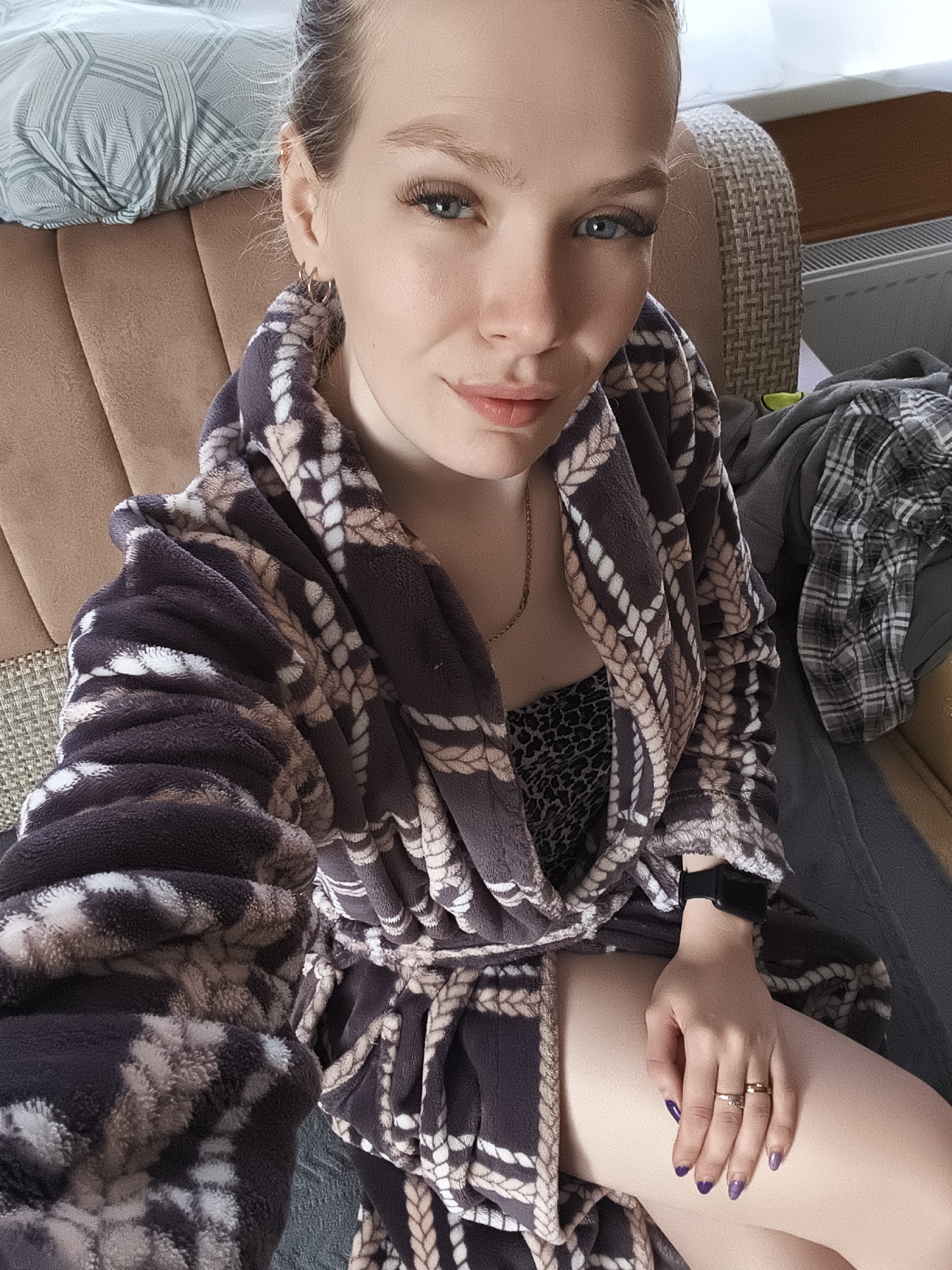 A fair-skinned woman with blue eyes and blonde hair sits in a brown chair. She wears a dark purple, rope-patterned bathrobe, and a leopard print top is visible underneath. She also wears a gold chain and has two gold rings on her left hand. Her nails are painted a dark purple. A black smartwatch is on her left wrist. To her right is a gray blanket and a plaid shirt. A pillow with a geometric pattern rests behind her head. A radiator is visible behind the chair. The location is possibly Русский Вожой, Russia.