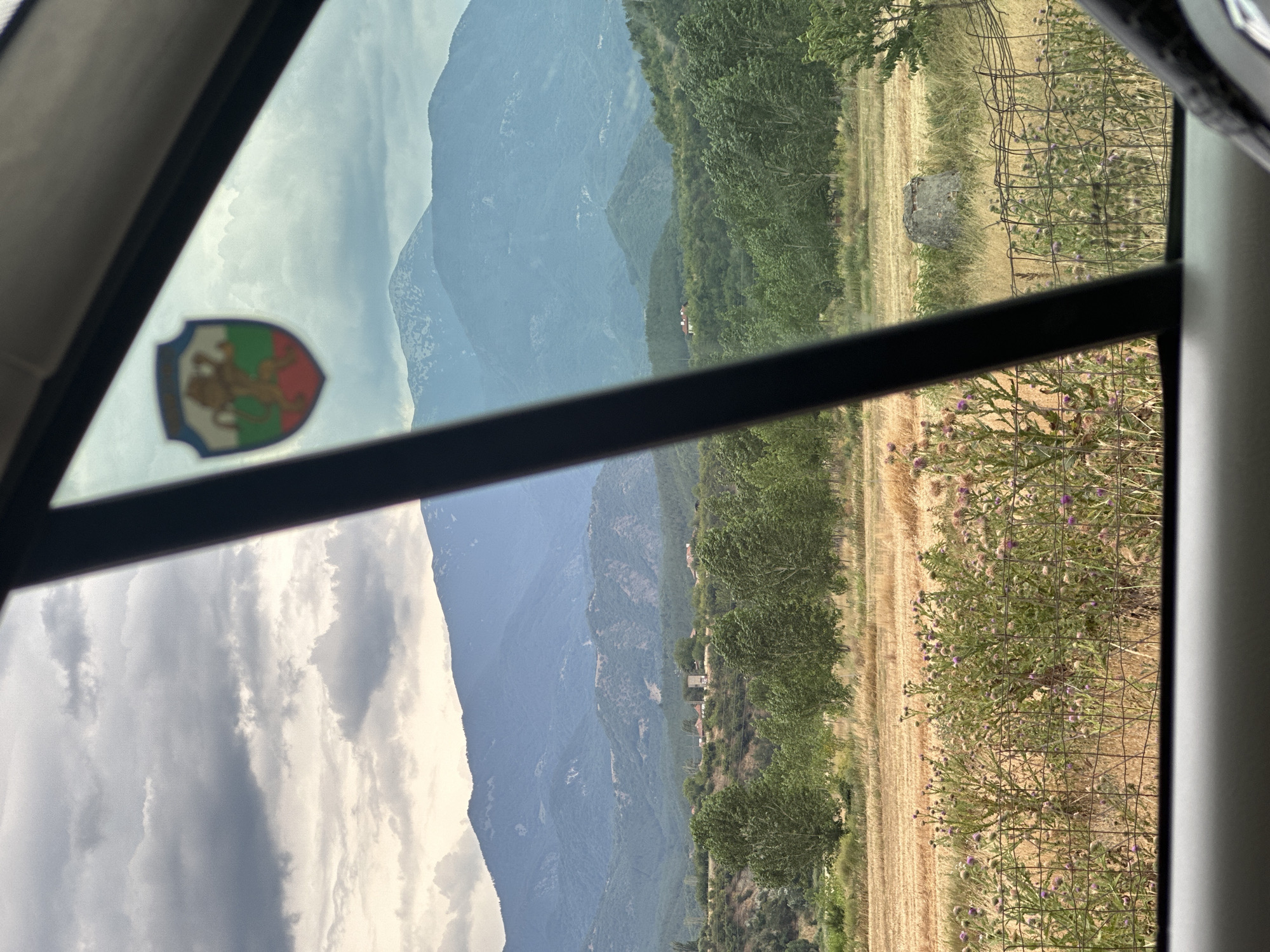The image is a reflection in a window, showing a landscape scene. The scene features layered mountains in the background, with trees and fields in the midground and foreground. The mountains appear to be covered in a light haze, suggesting a warm and possibly humid day. The midground is filled with lush green trees, interspersed with some smaller structures that appear to be houses or buildings. In the foreground, there is a field of dry, golden grass, possibly indicating a harvest season or a dry climate. There are also patches of wild vegetation with purple flowers and a wire fence running through them. A large, grey rock is visible in the field.
The reflection also shows a part of the interior of a vehicle, with the car window frame prominently visible. On the glass, a colorful shield-shaped sticker is affixed. This sticker displays a lion in the center, with red, green, and white sections. The overall impression is a journey through a scenic, mountainous region, likely in a rural area. The lighting suggests it is daytime, with the sun casting a gentle glow over the landscape. The presence of the sticker hints at a personal journey or connection to a specific place or group, possibly related to Bulgaria given the location context. There is no discernible text in the visible part of the image, other than what might be part of the sticker's design, which is not clear enough to read. The scene evokes a sense of travel and appreciation for natural beauty.