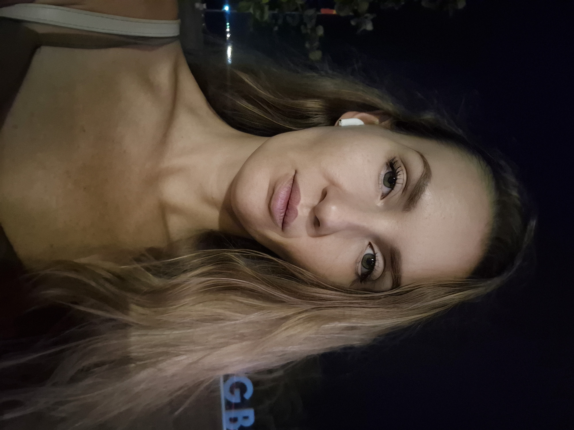 An outdoor, nighttime, close-up selfie shows a woman with long, blonde hair and green eyes. She is wearing a light-colored strap over her shoulder and a white earbud in her left ear. Her lips are slightly parted and her expression is neutral. In the background, out of focus, are some trees and a sign with large blue letters spelling "GB". The setting appears to be an urban area in Đà Nẵng, Vietnam, possibly near a waterfront or entertainment district.