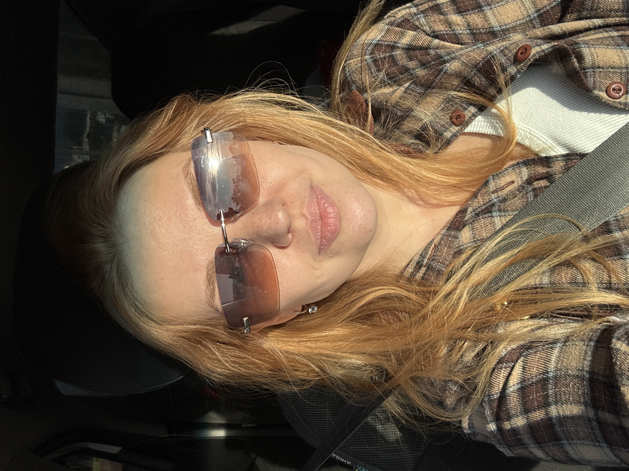 A person with light reddish-blonde hair and sunglasses is in the passenger seat of a car. The person is wearing a brown plaid shirt over a white top and a seatbelt. The car interior is black. The reflection in the sunglasses shows an outdoor environment. The location of Podosenikha, Russia is not possible to be ascertained from this image.