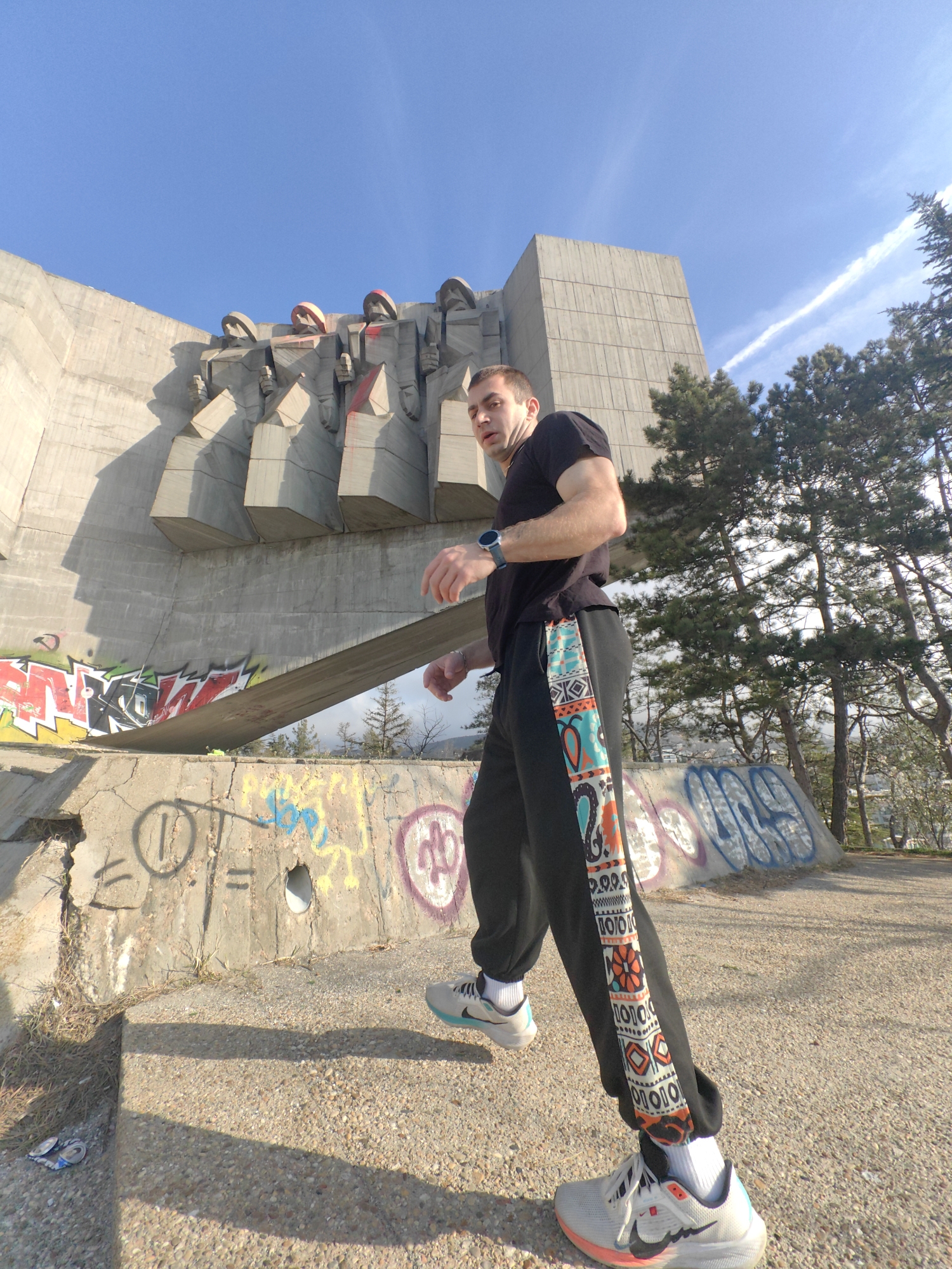 The image depicts a male individual walking on a light-colored gravel path in front of the Monument to the Bulgarian-Soviet Friendship in Varna, Bulgaria. The monument, a large brutalist concrete structure, dominates the left and center background, featuring stylized human figures on its facade, some of which are defaced with red spray paint. The lower concrete walls of the monument and its base are extensively covered in multi-colored graffiti tags.
The male subject, positioned in the lower center and right, is captured in mid-stride, facing slightly left with his gaze directed toward the camera. He has short dark hair and is wearing a black short-sleeved t-shirt, black sweatpants with a prominent vertical, multi-colored geometric pattern stripe on the outer legs, white athletic socks, and white and gray athletic shoes with a black Nike swoosh and orange/light blue accents. A blue smartwatch is visible on his left wrist. His arms are slightly bent, consistent with a walking motion.
The ground surface is concrete with visible gravel aggregate. Shadows are cast to the left foreground. Two crushed aluminum cans are present on the ground in the lower-left corner. Pine trees and sparse vegetation are visible in the right and distant background. The sky is clear and blue, with a faint white contrail visible in the upper right.