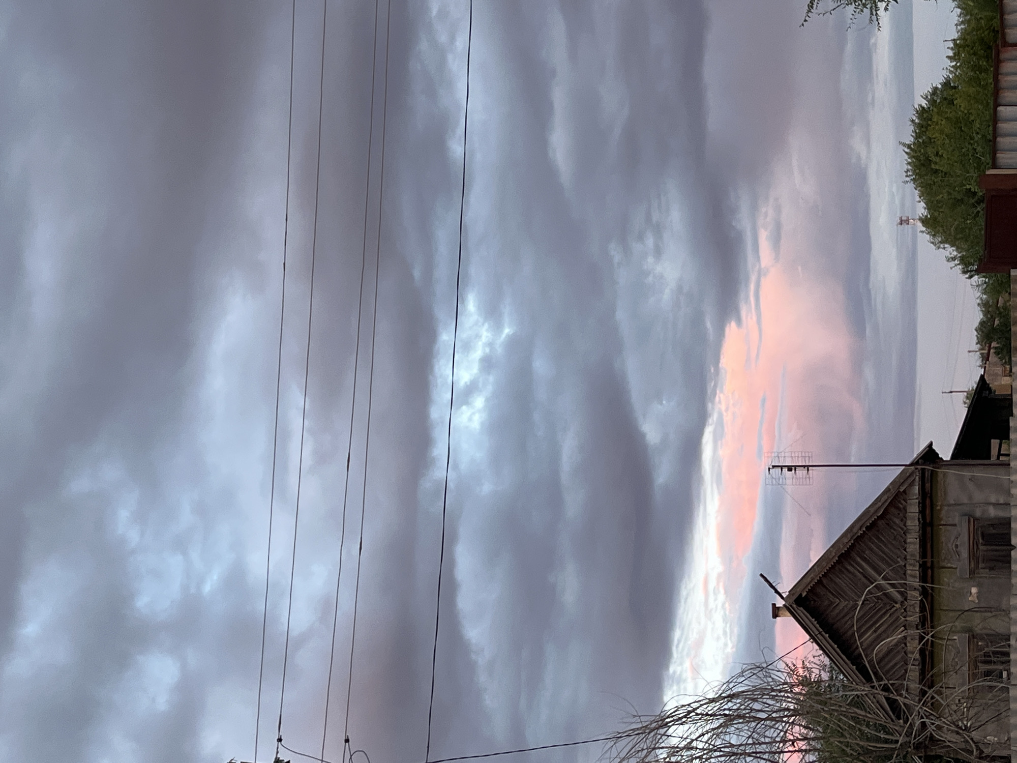 This media file captures a twilight sky over a rural scene in Dubovka, Russia. The sky is filled with dramatic, swirling clouds, some dark and ominous, others tinged with soft pink and orange hues, suggesting the last moments of sunset. Several thin, dark power lines stretch across the frame from left to right, bisecting the clouds.
In the foreground, the dilapidated roof of a wooden house is visible, its weathered shingles and a small chimney indicating age and disuse. An old television antenna juts out from the peak of the roof. To the right, a dense, green tree offers a natural contrast to the man-made structures and the dramatic sky. Bare, thorny branches of a bush or tree in the lower foreground partially obscure the view of the house and the sky.
The overall mood is one of serene melancholy, amplified by the fading light and the worn appearance of the house. The scene suggests a quiet evening in a rural area, with the natural beauty of the sky juxtaposed against the signs of rural life. There is no visible text in the media file.