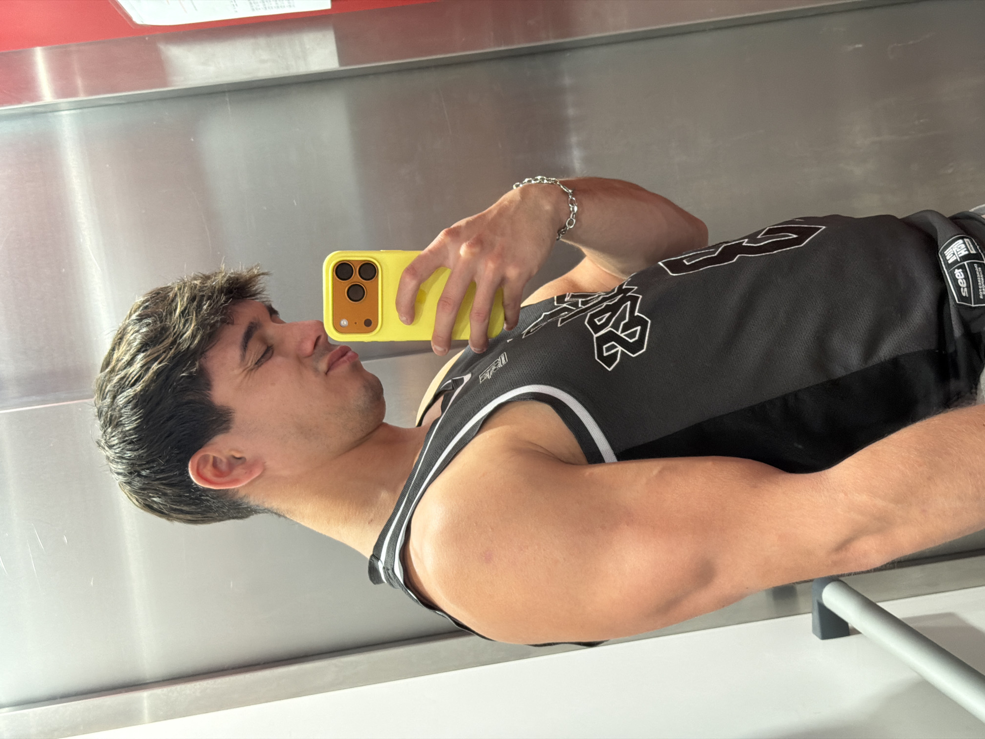 A young man with short, dark, slightly messy hair is captured in a mirror selfie, rotated 90 degrees clockwise. He appears to be standing, reflected in a large, polished metal surface that forms the background, with a red stripe visible at the top edge of the reflection and a grey horizontal bar further down. His eyes are closed, and his lips are slightly pursed, suggesting a posed or playful expression.
He wears a black sleeveless basketball jersey with white trim and prominent white graphics, including the number "23." A small fabric tag on his right shoulder shows partial text, possibly including "NEW YORK" and "ESSENTIALS," along with smaller, illegible words. A silver chain bracelet is visible on his right wrist, which holds a bright yellow smartphone featuring a triple-lens camera array. The man's athletic build is visible, particularly his toned arms. The setting, an indoor space with reflective surfaces, offers no specific visual cues to its location within Sofia, Bulgaria. The time of day is not discernible.